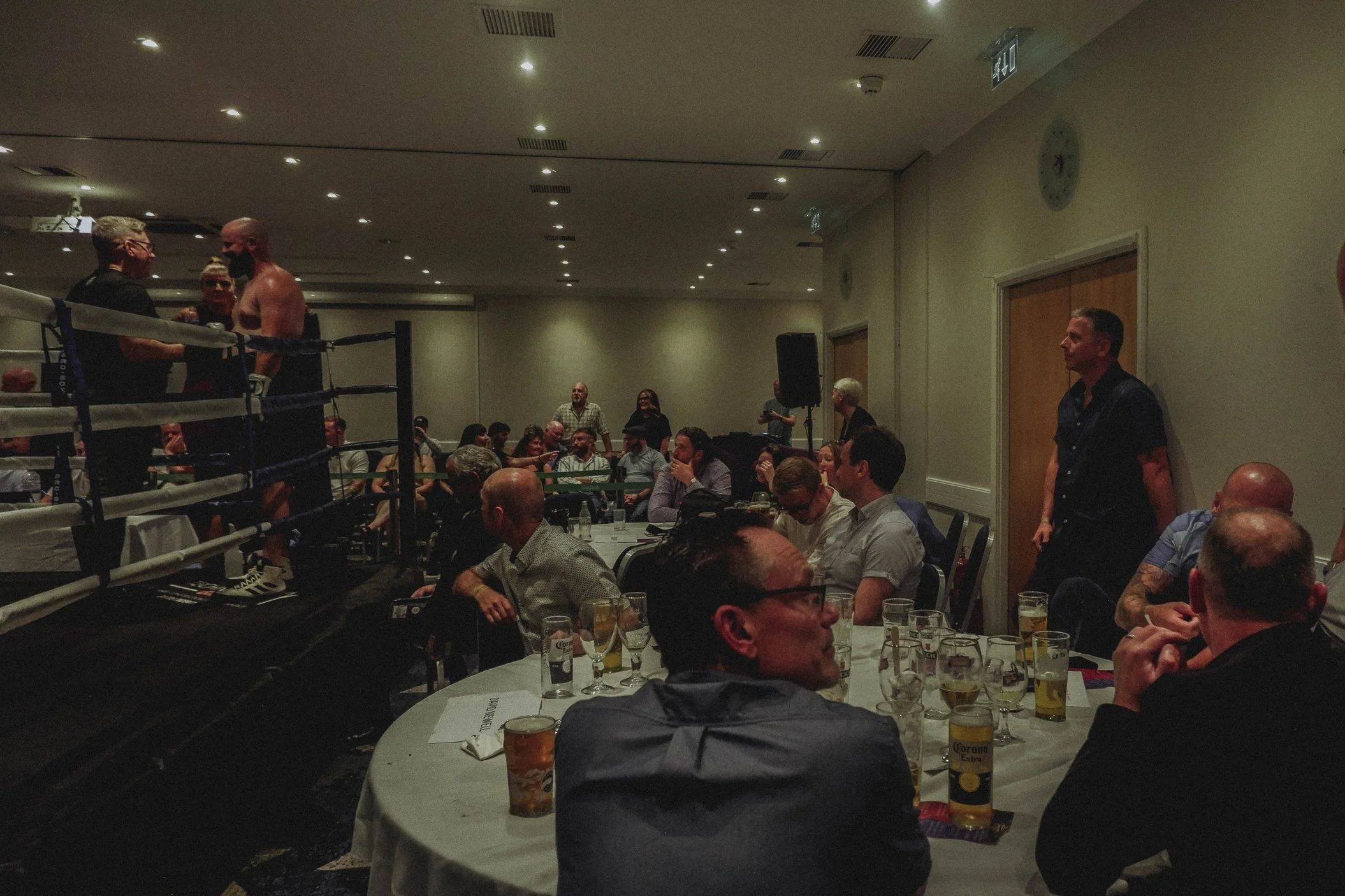 A boxing event in an indoor venue with a ring on the left and seated audience watching, some standing near the ring.