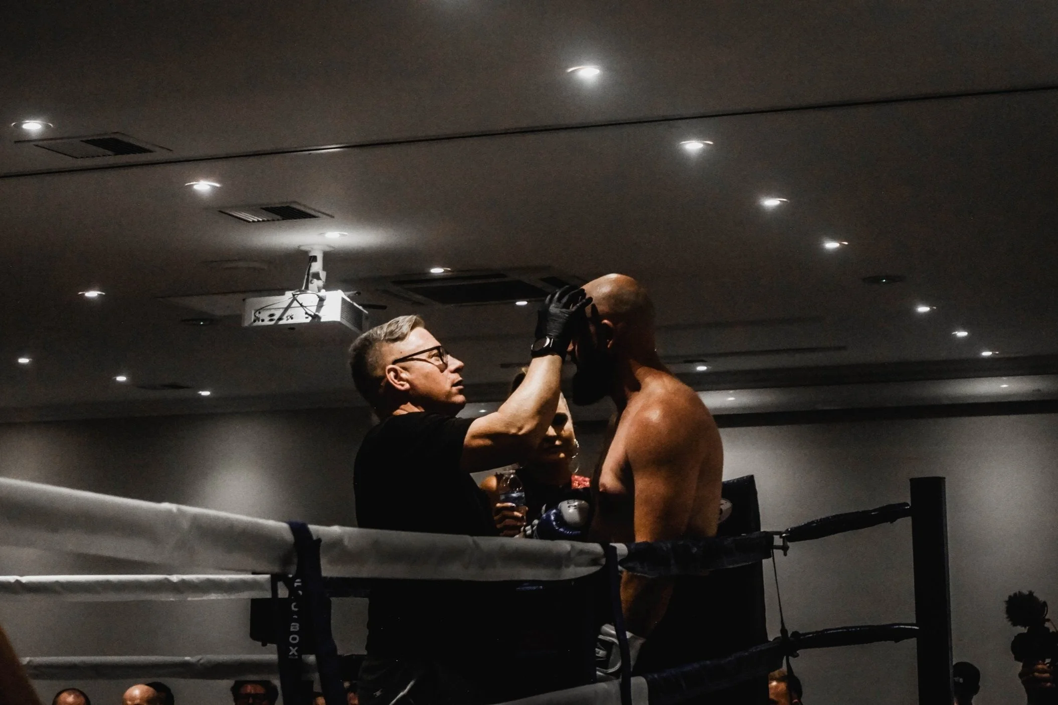 A boxing referee attends to a shirtless boxer in a boxing ring during a match, with the referee adjusting his headgear.