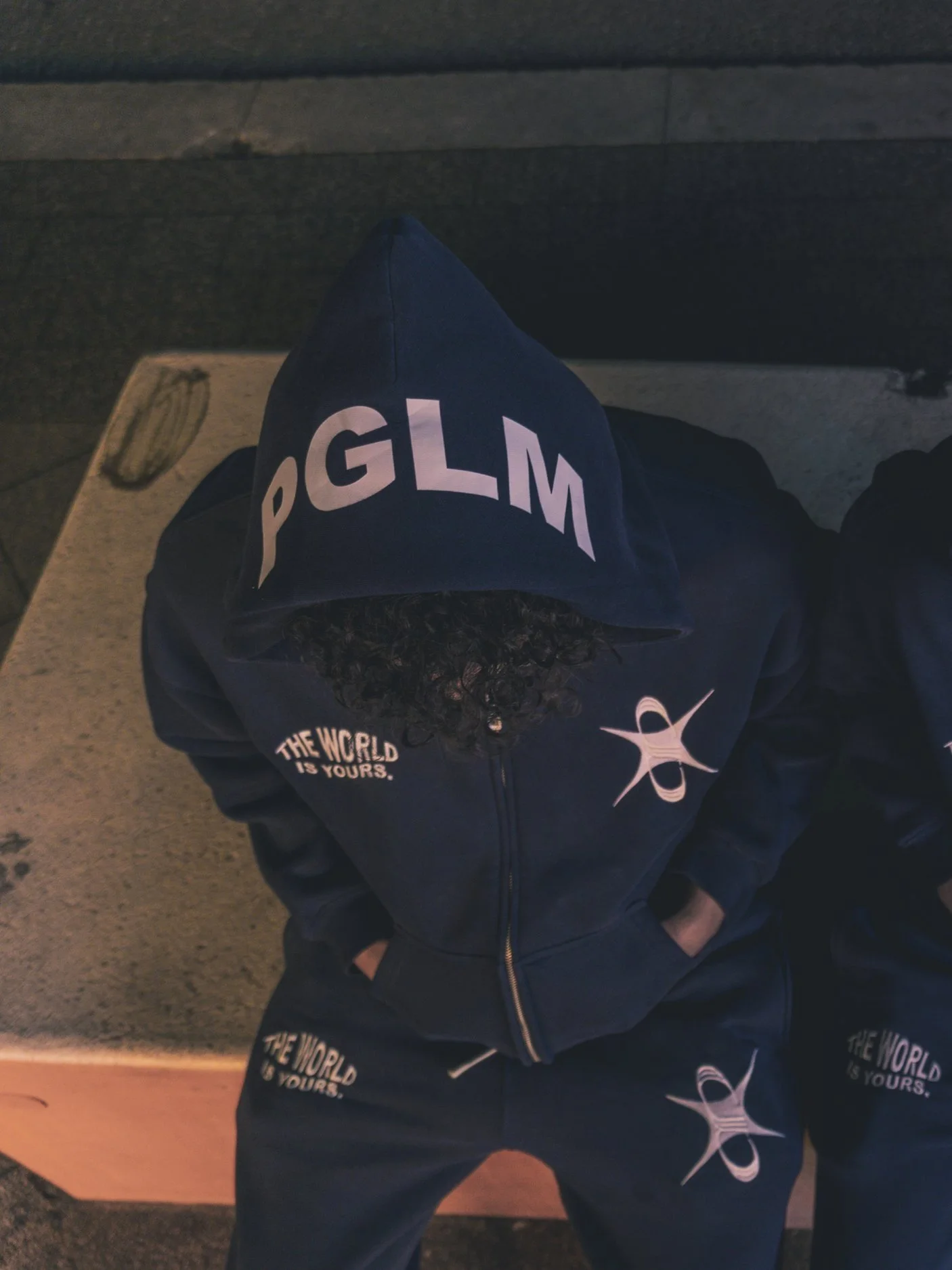 A person wearing a dark hoodie with the word 'PGLM' on the hood and 'THE WORLD IS YOURS' on the shirt, standing with hands in pockets, with dark curly hair visible under the hood, in a nighttime outdoor setting.