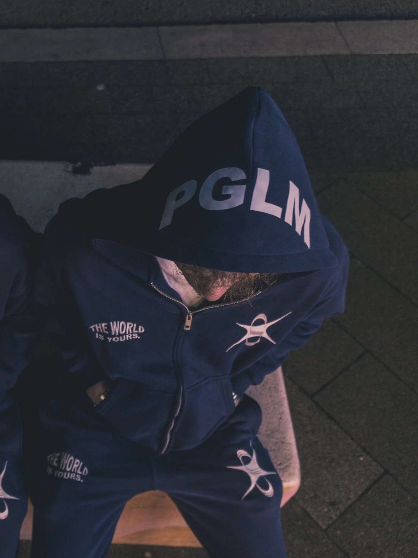 Individual sitting outdoors wearing a navy blue hoodie and sweatpants, with the words 'THE WORLD IS YOURS' and a logo printed on both, on dark pavement, seen from above with the hood up.