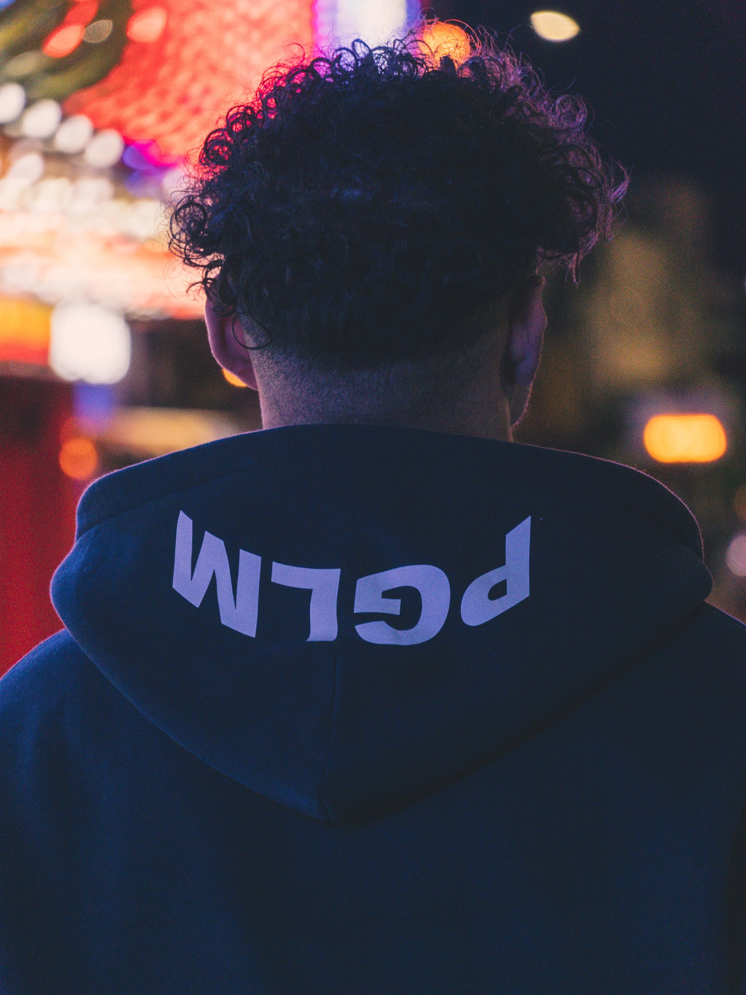 Back view of a person with curly hair wearing a dark hoodie with 'PLDM' printed upside down on the hood, standing in a city at night with colorful blurred lights.