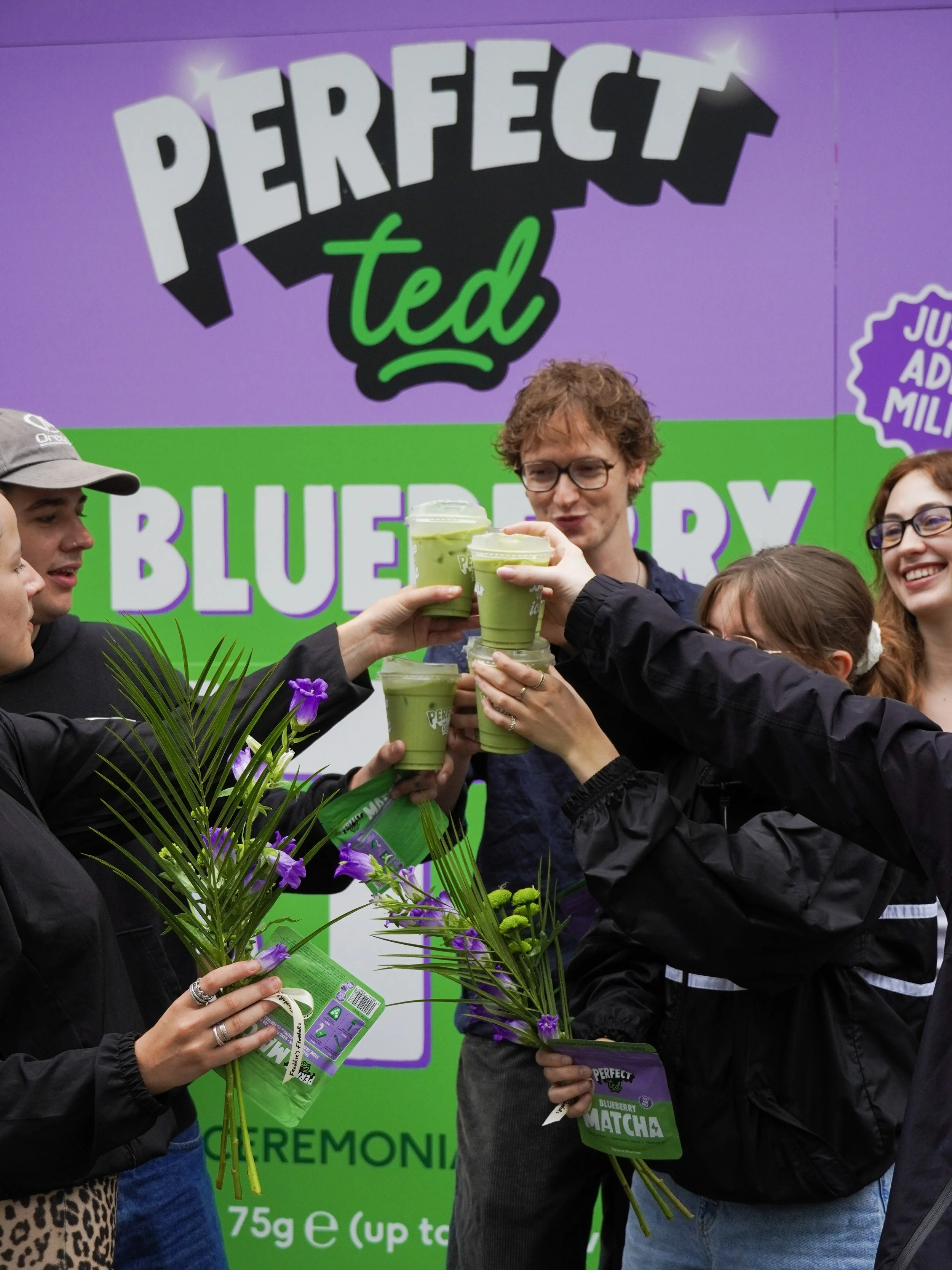 Group of people celebrating with matcha drinks and purple flowers in front of a purple and green promotional banner for Perfect Ted blueberry matcha product.