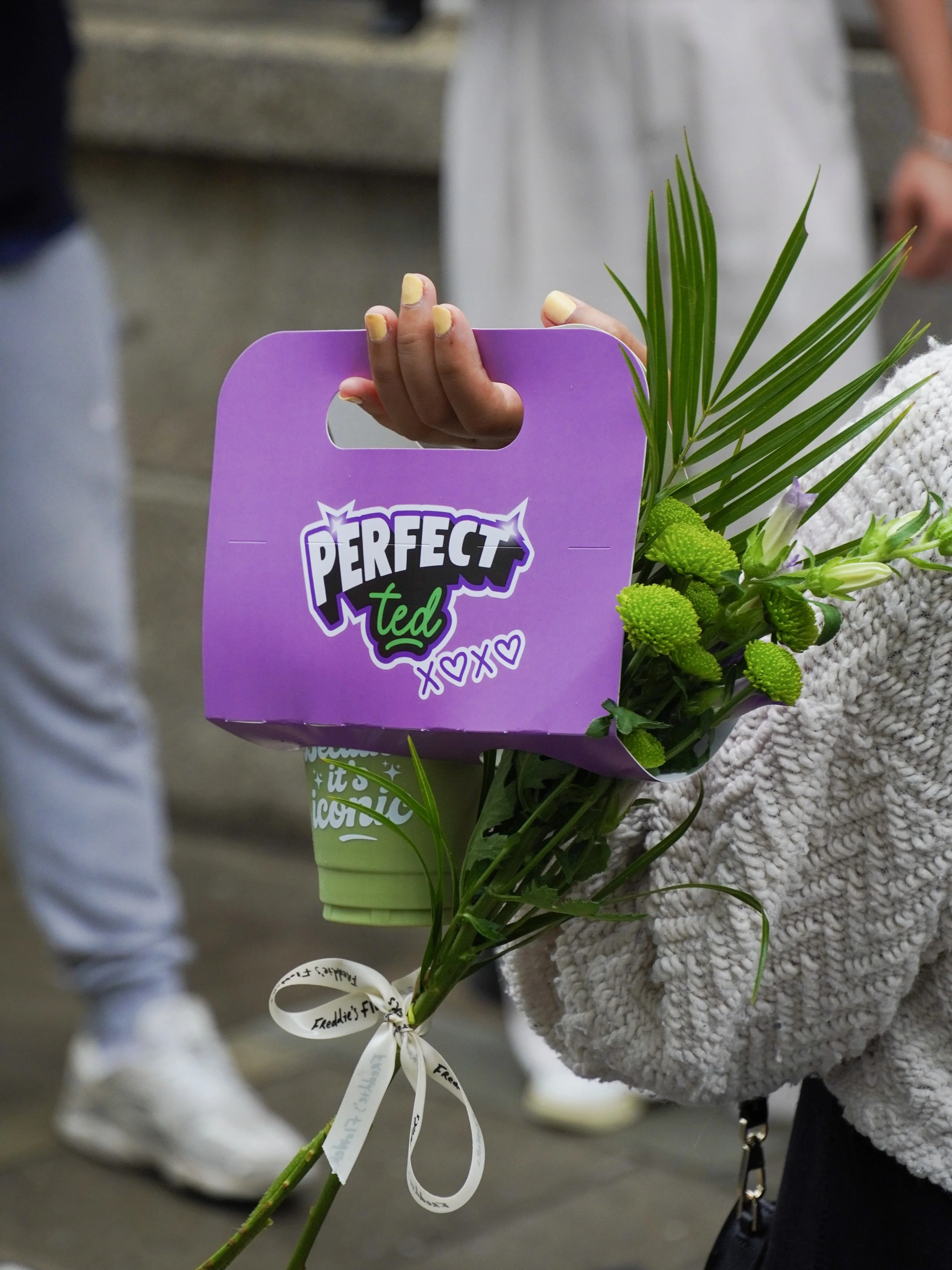 Person holding a purple gift bag with a sticker that says 'PERFECT ted' and small hearts, attached to a bouquet of green flowers and leaves.