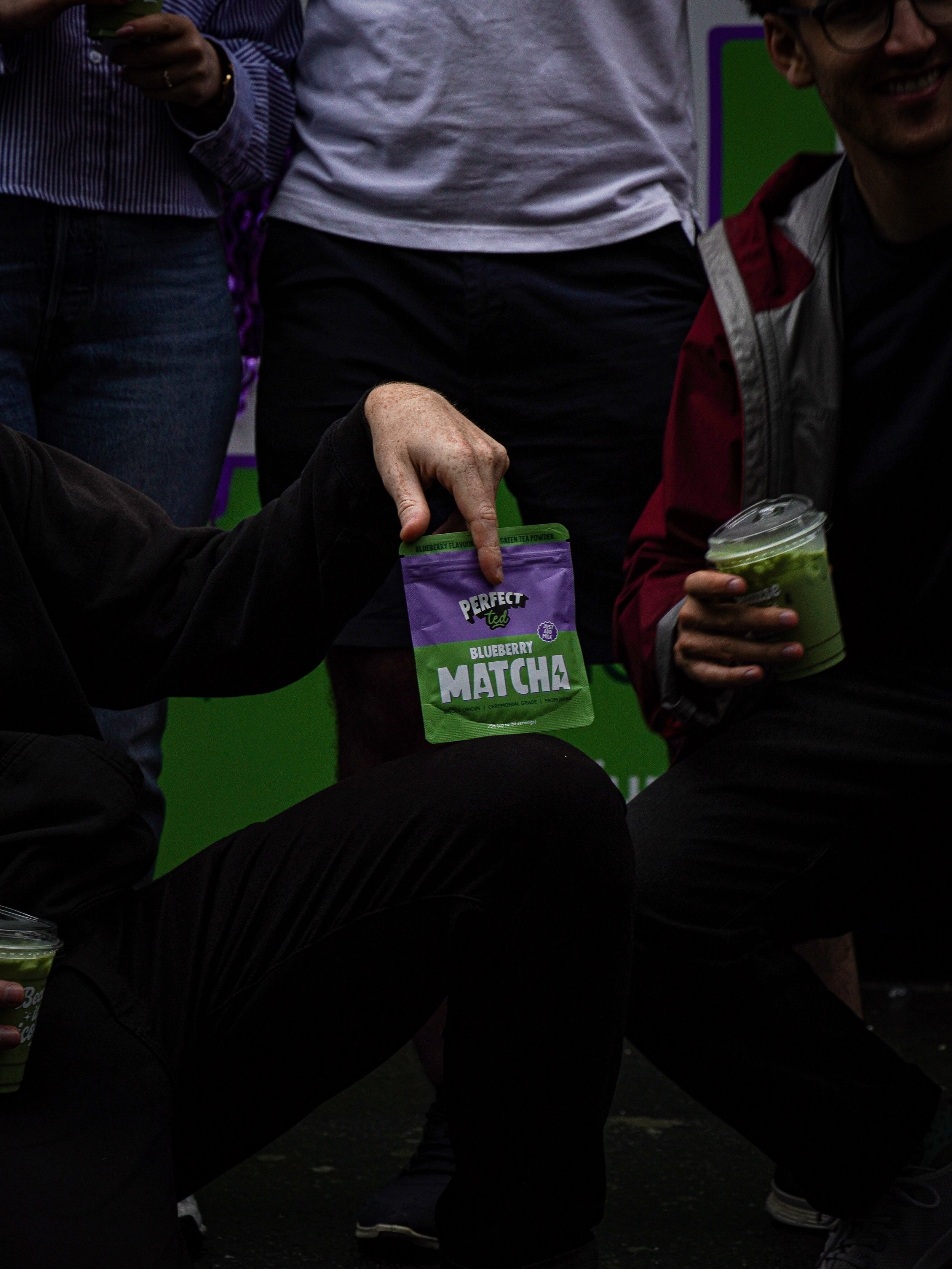 People holding packets of Perfect Tea Blueberry Matcha and cups of green tea latte at a promotional event.