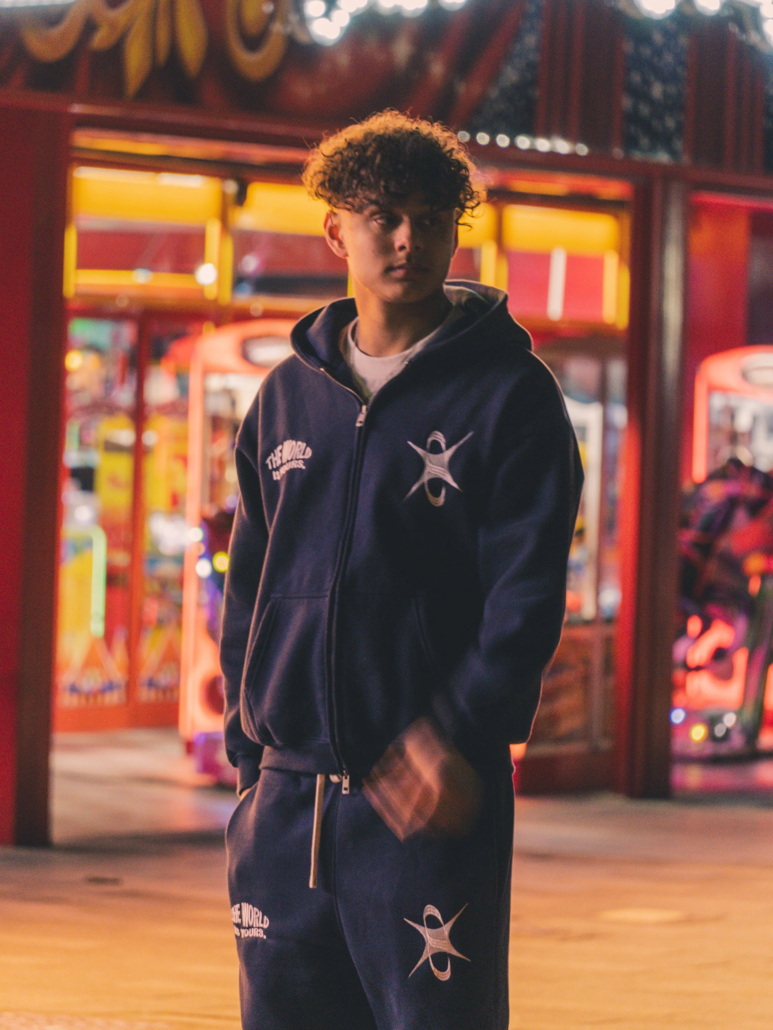 A young man with curly hair stands outside an arcade at night, wearing a navy hoodie and sweatpants with white logos and text, in front of brightly lit arcade machines.