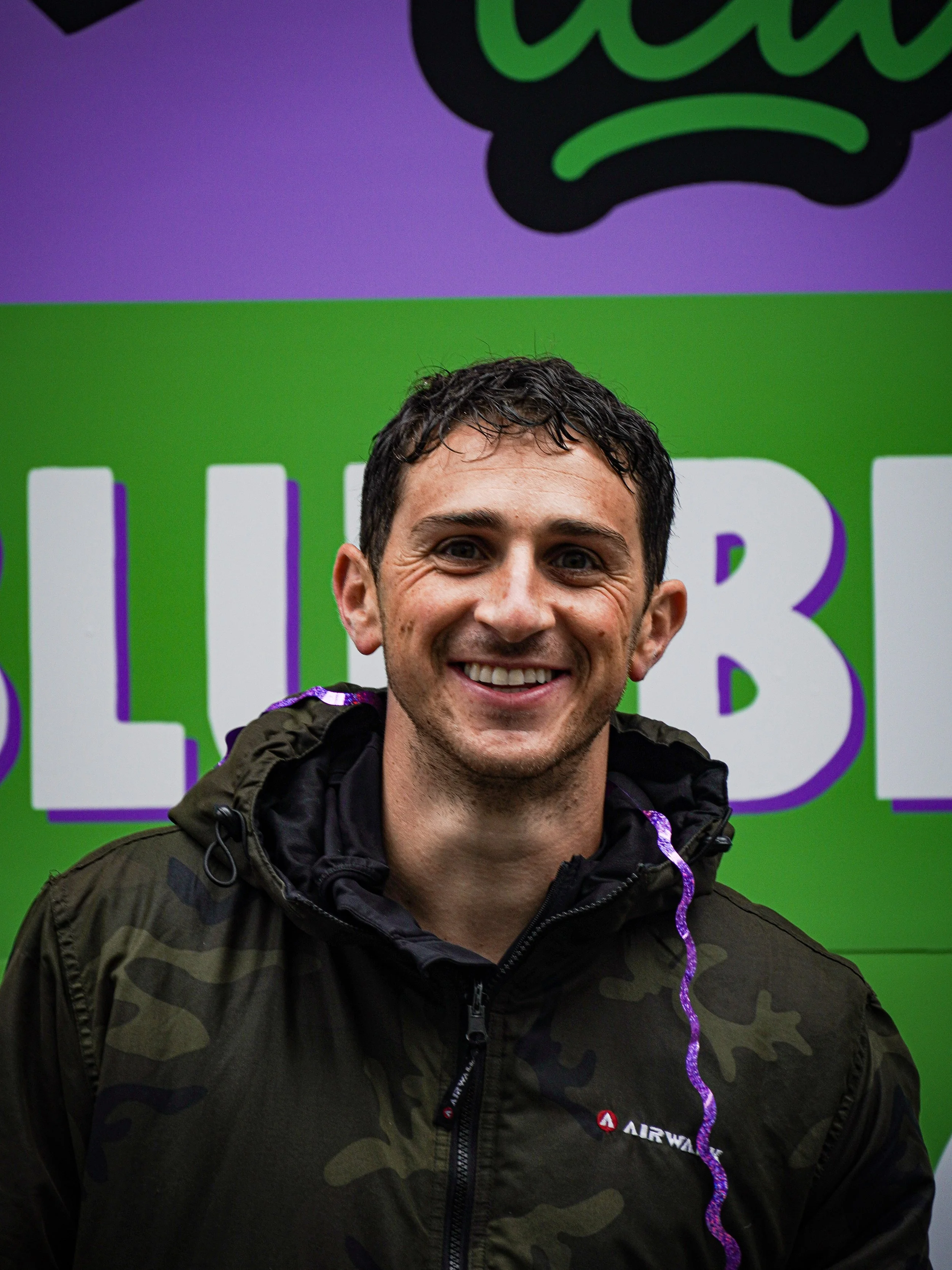 A smiling man with short dark hair wearing a camouflage jacket standing in front of a brightly colored background with large text.
