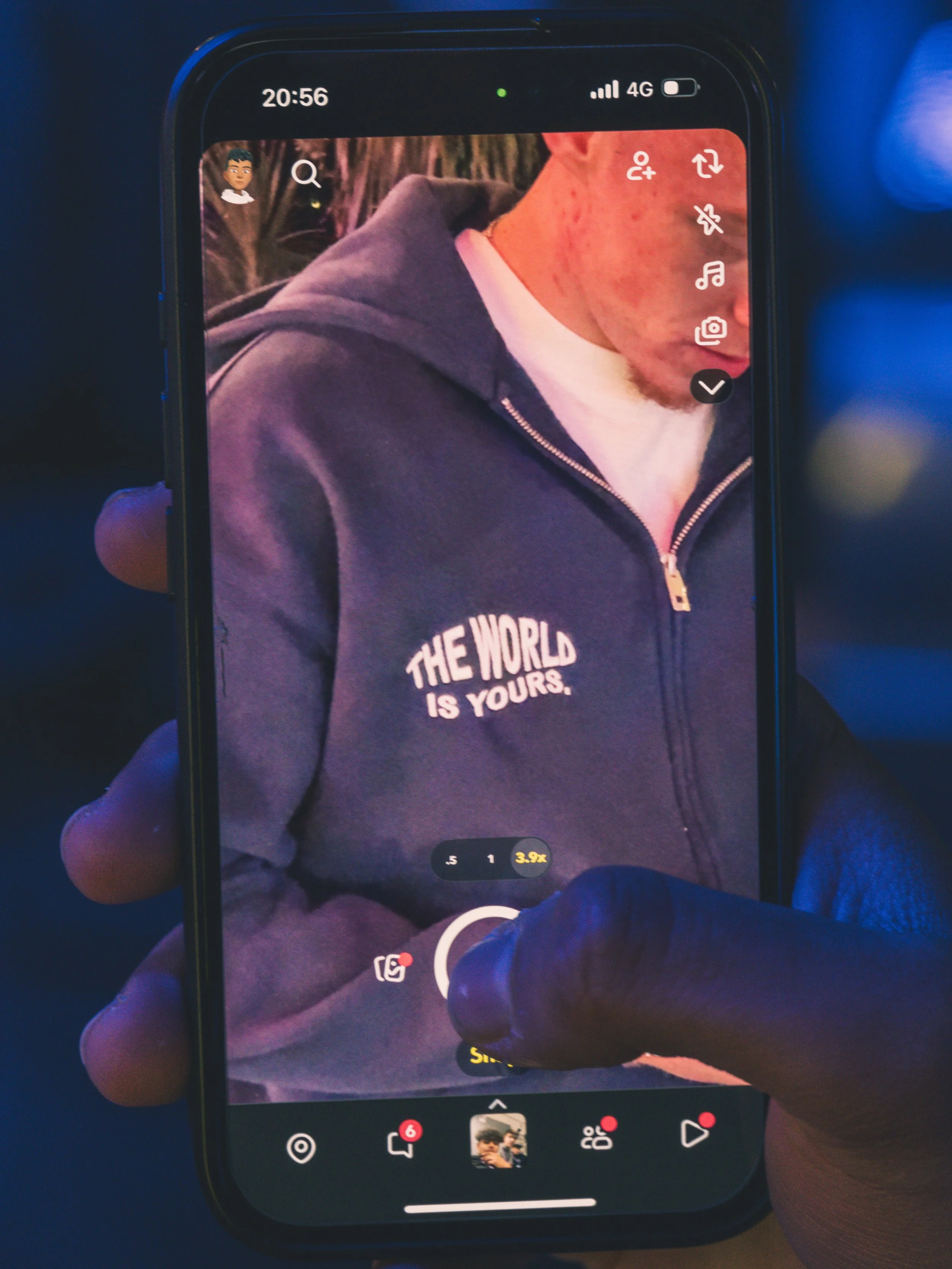 Close-up of a smartphone screen showing a video recording of a person wearing a navy hoodie with the text 'THE WORLD IS YOURS' on it. The person's face is partially visible, looking down. The app interface displays various icons and a red record butt