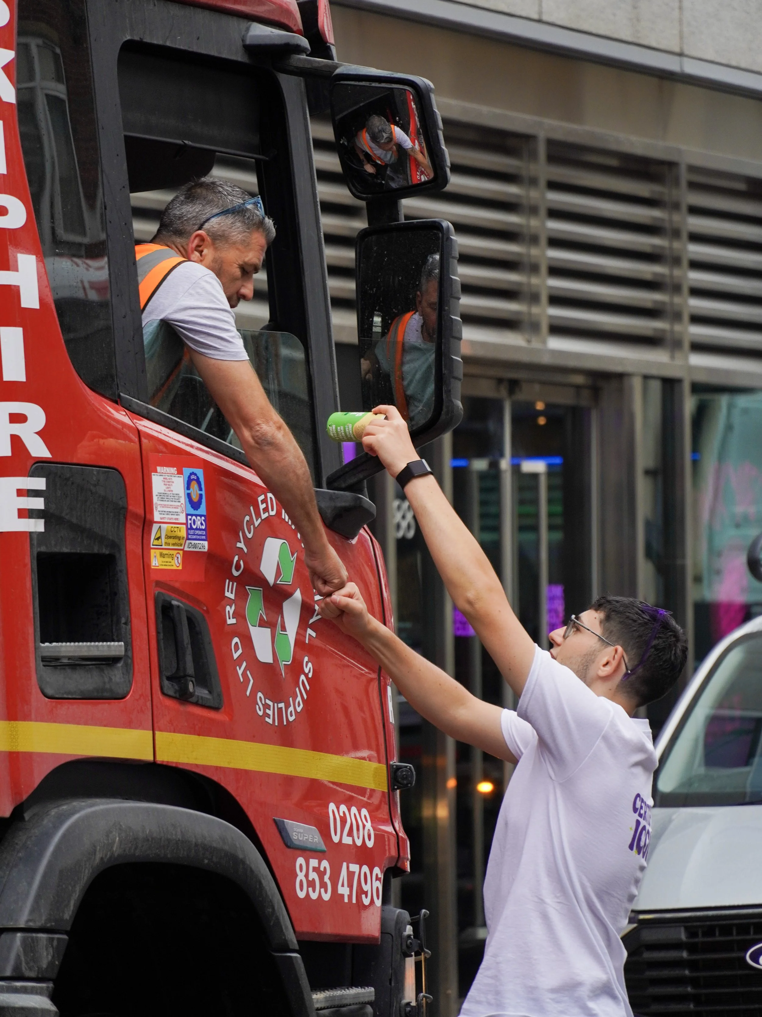 A person in a white shirt handing a bottle of a green beverage to a firefighter in the cab of a red fire truck, with the firefighter reaching out to receive it.
