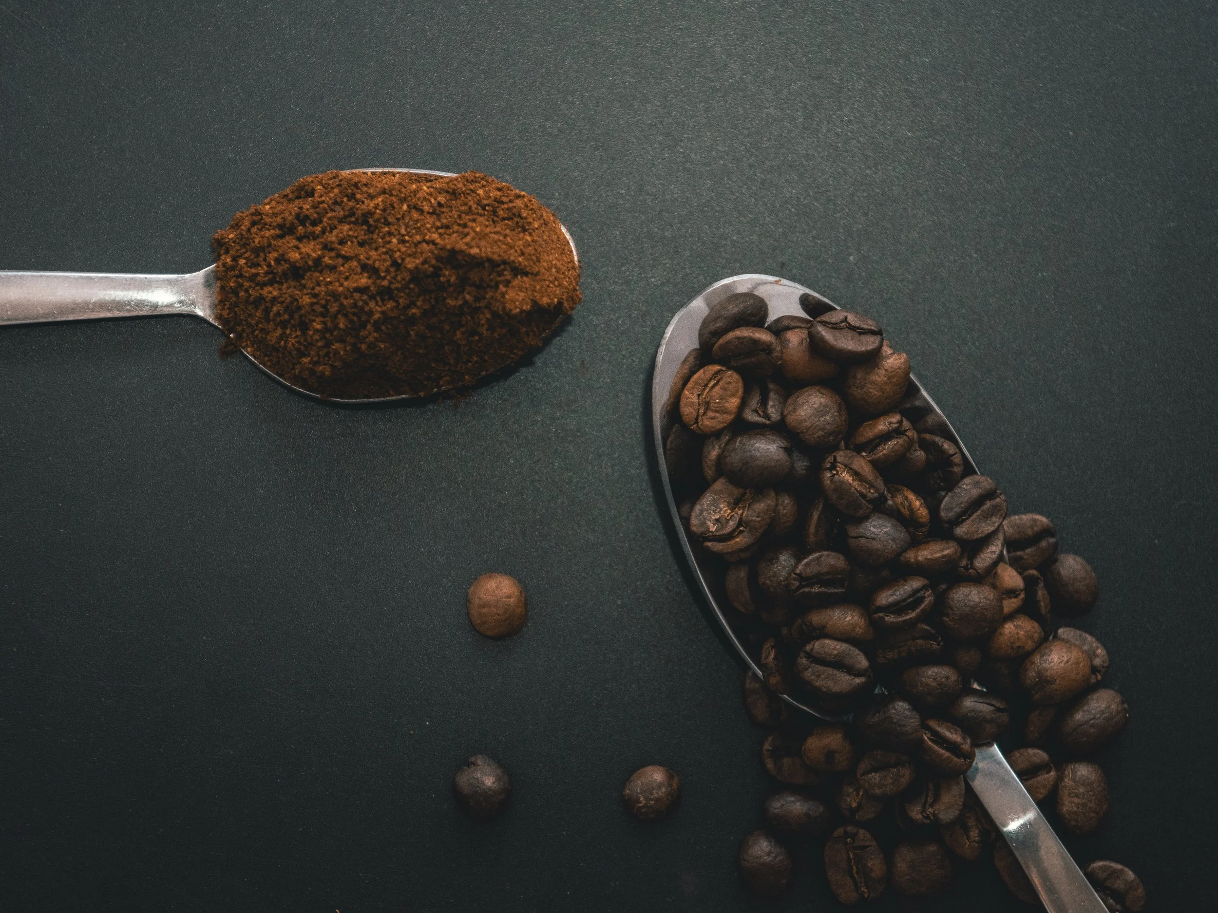 A metal spoon filled with ground coffee, a metal scoop filled with whole coffee beans, and a few loose coffee beans on a dark surface.