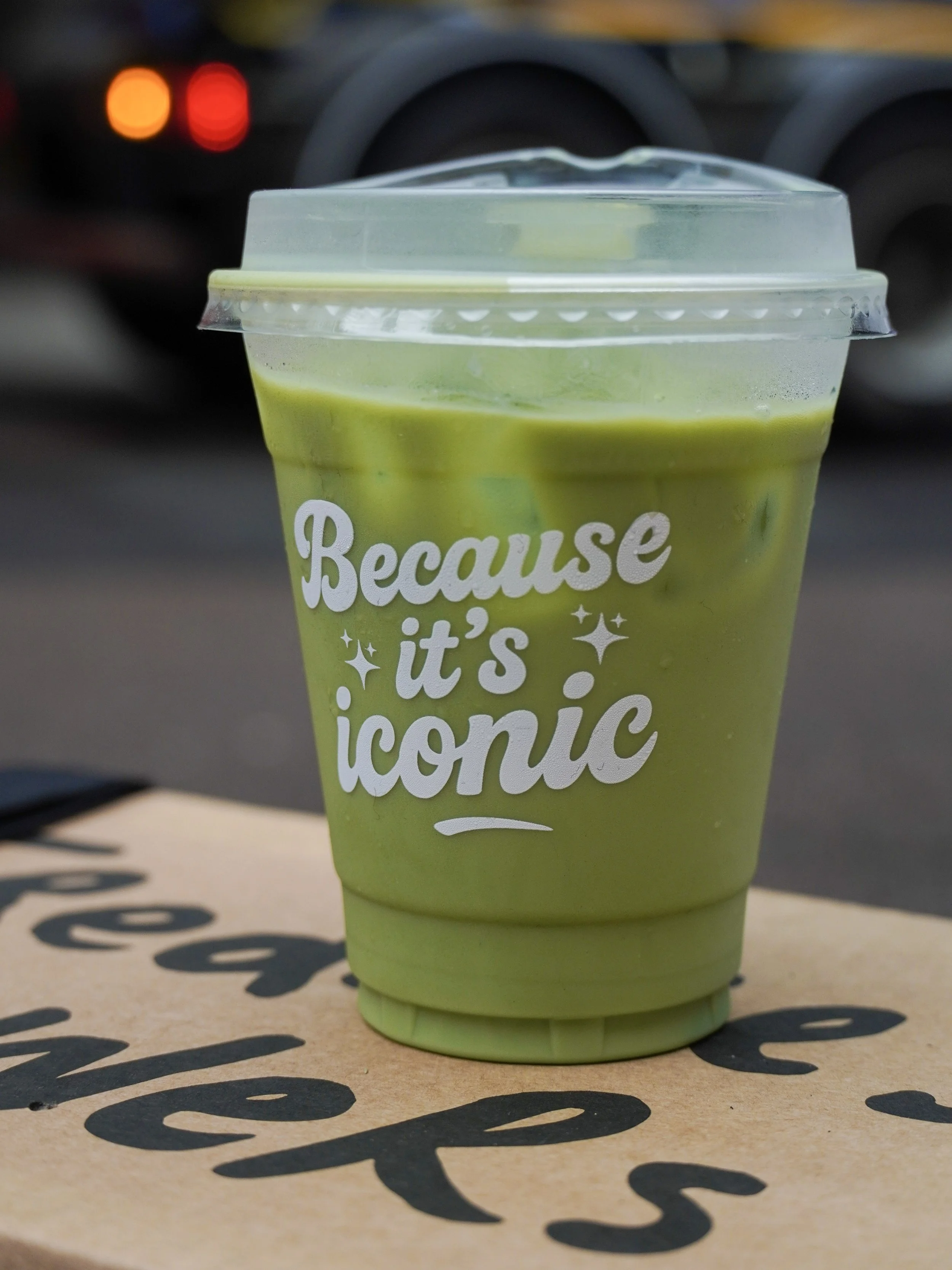 Green iced beverage in a clear plastic cup with a dome lid, sitting on a cardboard box with black writing.