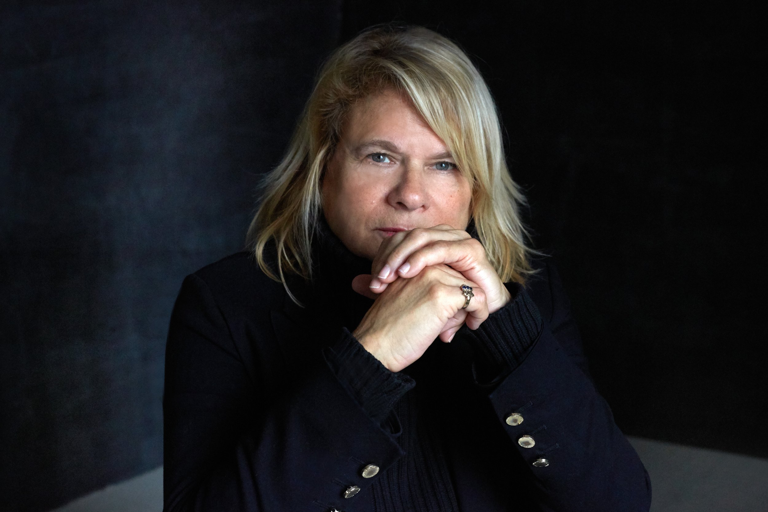 A person with blonde, wavy hair wearing a black leather jacket, resting their chin on clasped hands, looking intently at the camera against a black background.