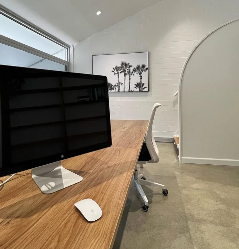 Modern office space with a wooden desk, an iMac computer, and a white office chair. A large window on the left side and a framed picture of palm trees on the white brick wall.