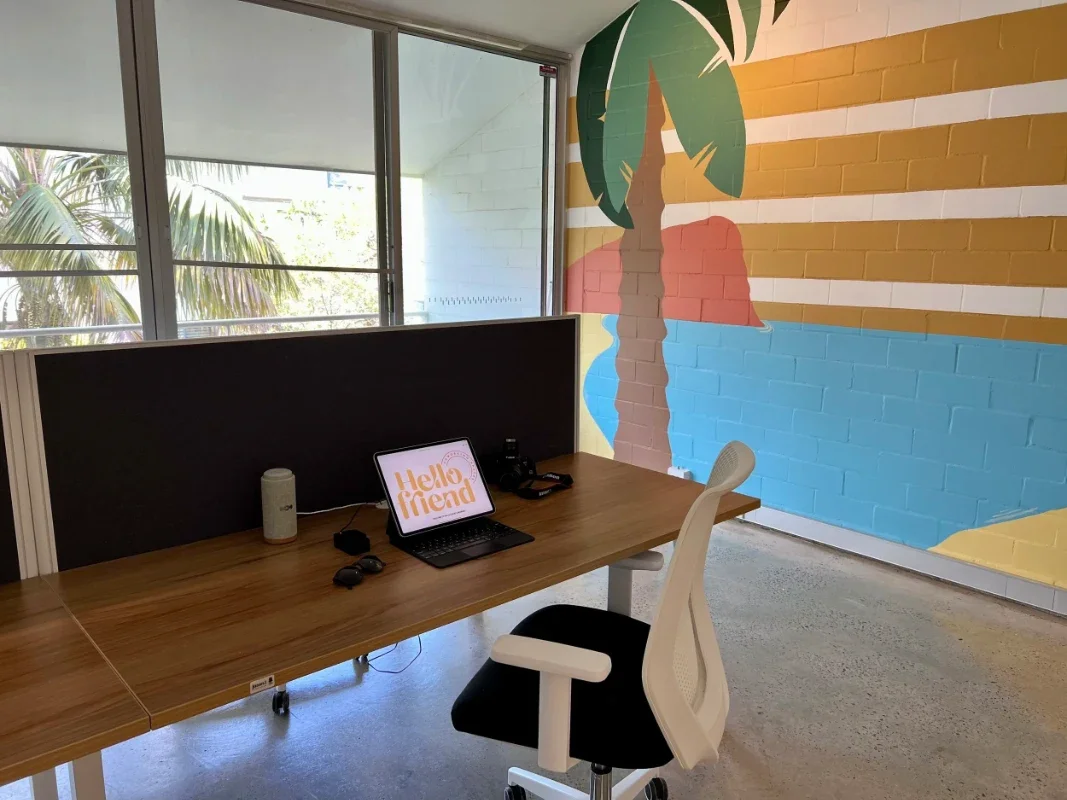 Office setting with a wooden desk, a white ergonomic chair, a computer monitor displaying 'Hello Friend', a camera, sunglasses, a speaker, and a large colorful mural of a palm tree, ocean, and cliffs on the brick wall.