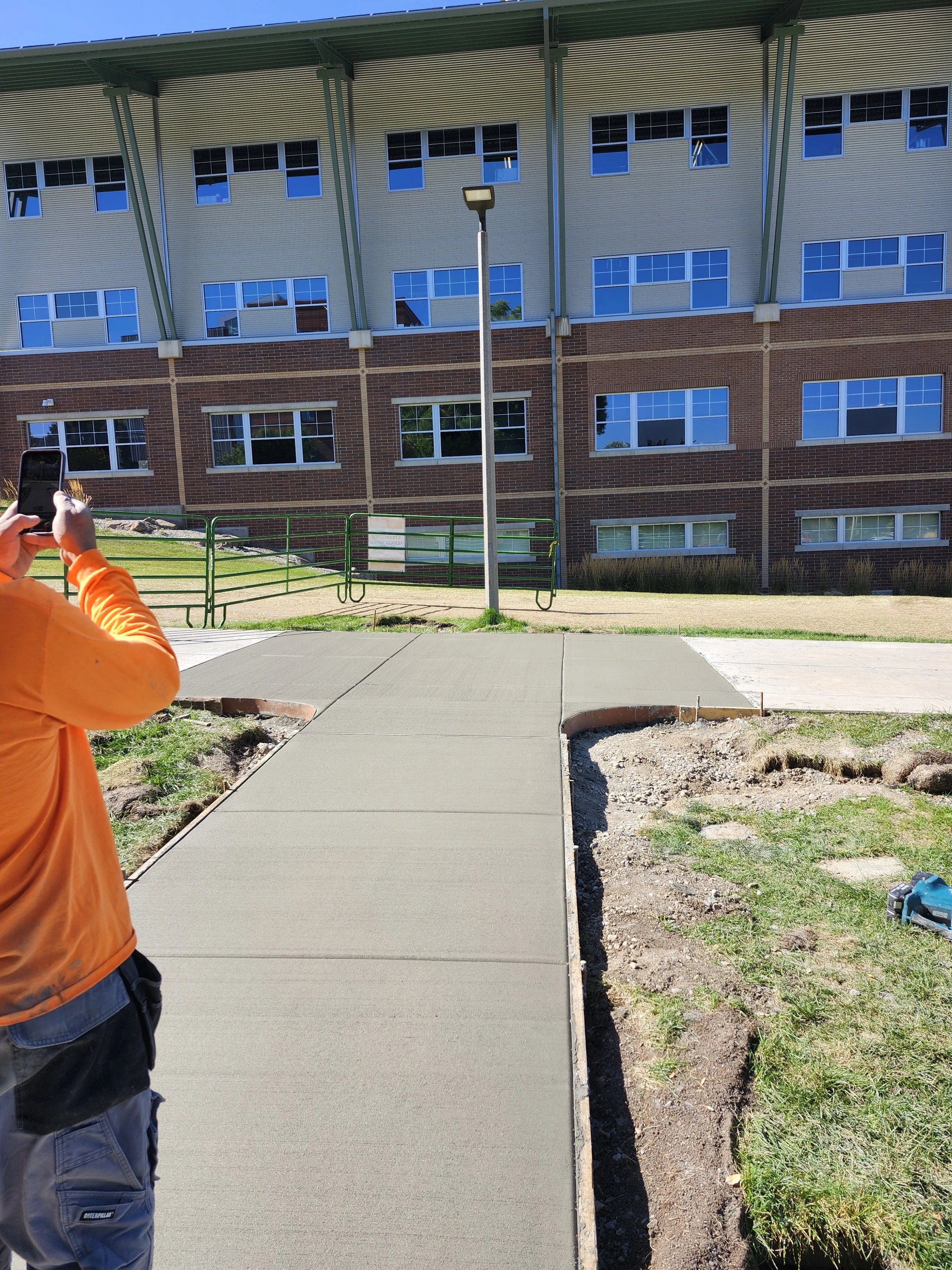 Person in orange jacket taking a photo of a new concrete sidewalk on a sunny day, with a modern building with large windows in the background.