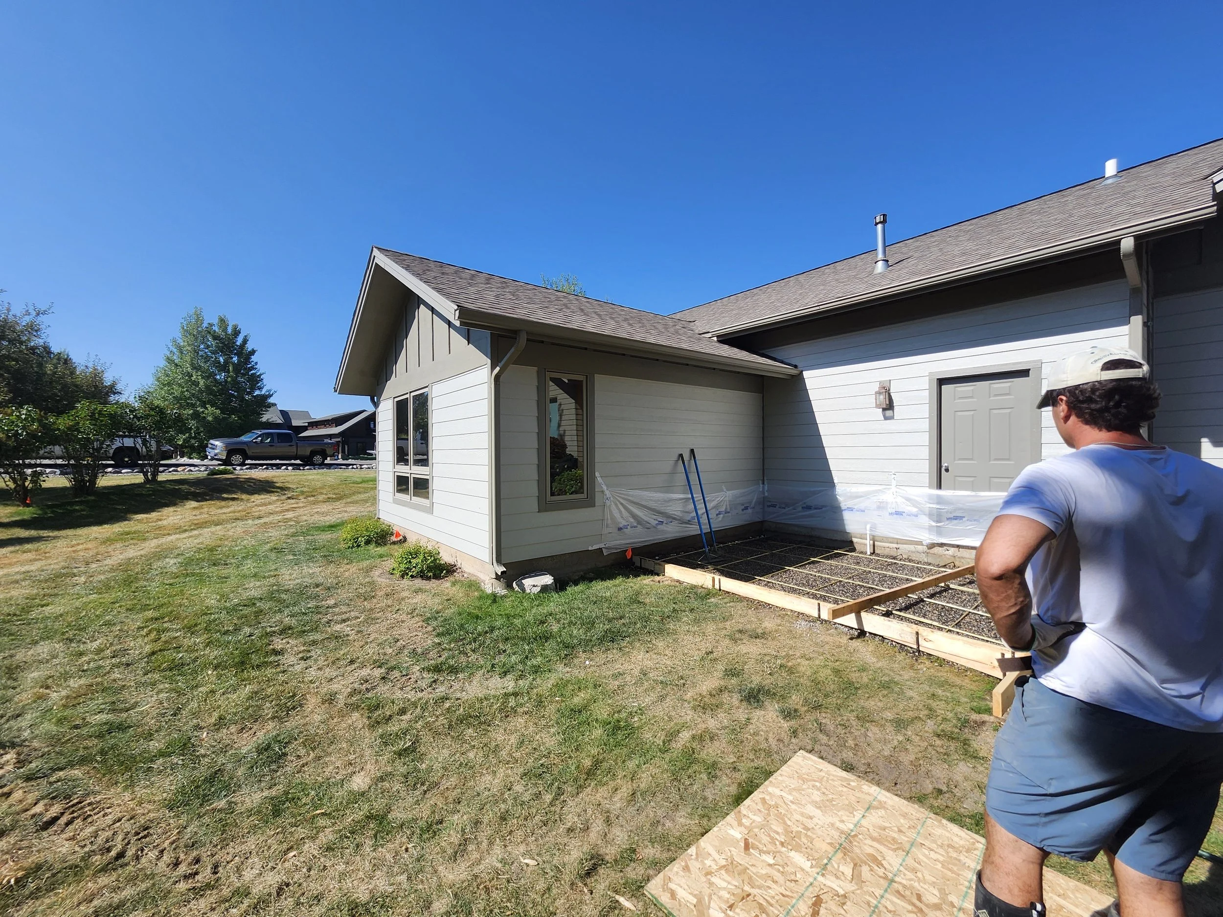 A person standing in a backyard, preparing a concrete foundation for a new patio or deck, with a partially constructed wooden frame and gravel base, next to a grey house under a clear blue sky.