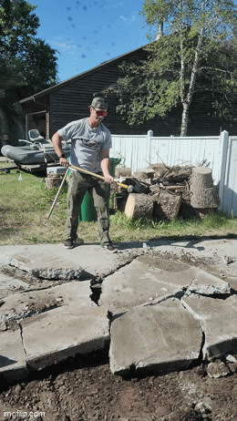 A man in a gray shirt and baseball cap is working on breaking up a concrete patio with a pickaxe. There is a white fence, a dark building, and trees in the background under a blue sky.