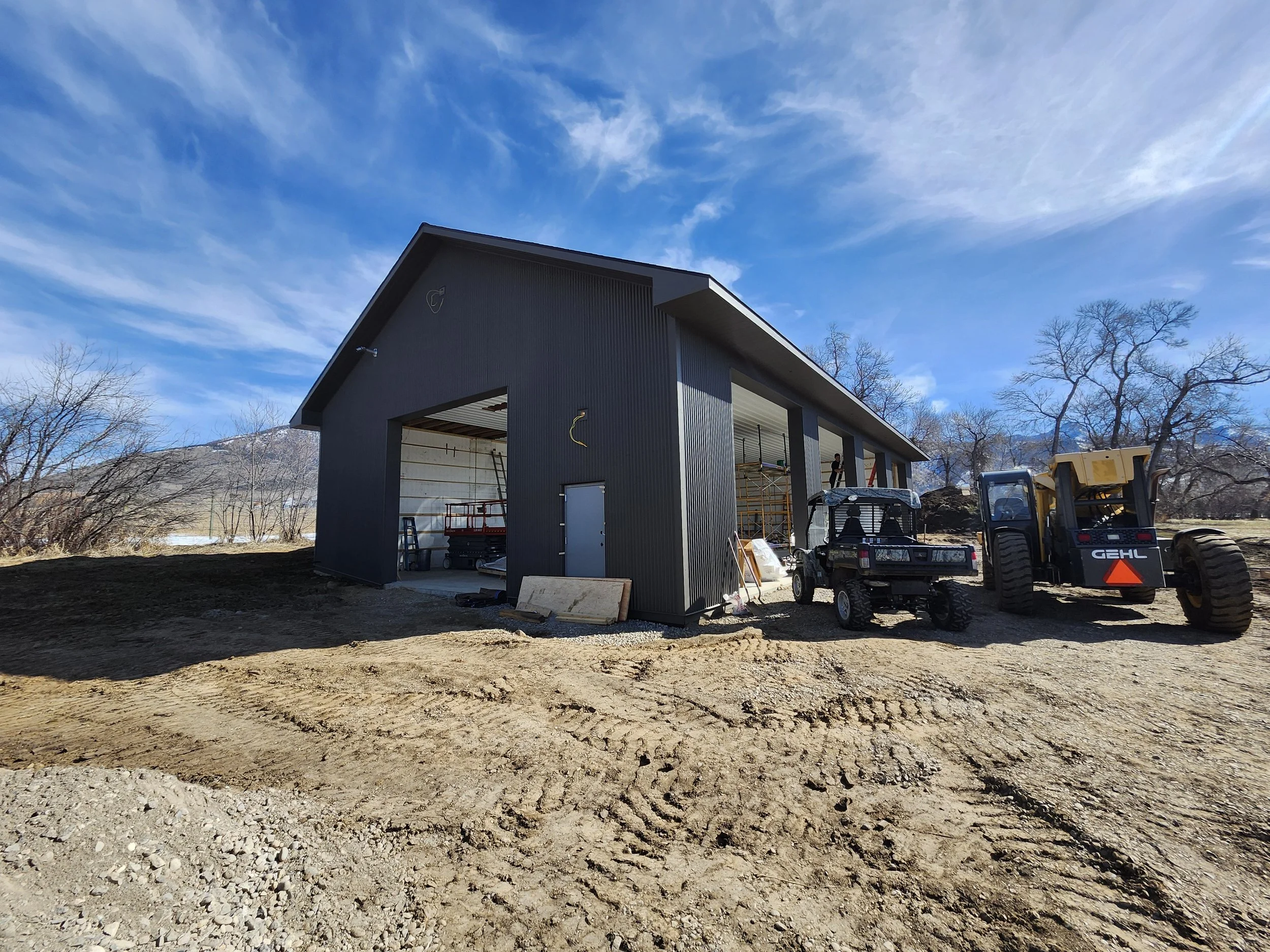 Construction site with a partially built dark gray metal building, construction vehicles including a small utility vehicle and a larger earth-moving machine, and a dirt ground under a blue sky with some clouds.