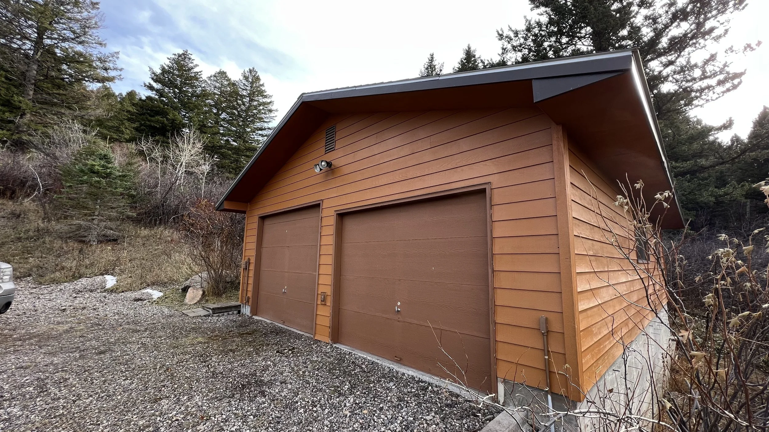 A wooden garage with two brown doors and a sloped roof, situated in a rural area among trees with a gravel driveway in front.