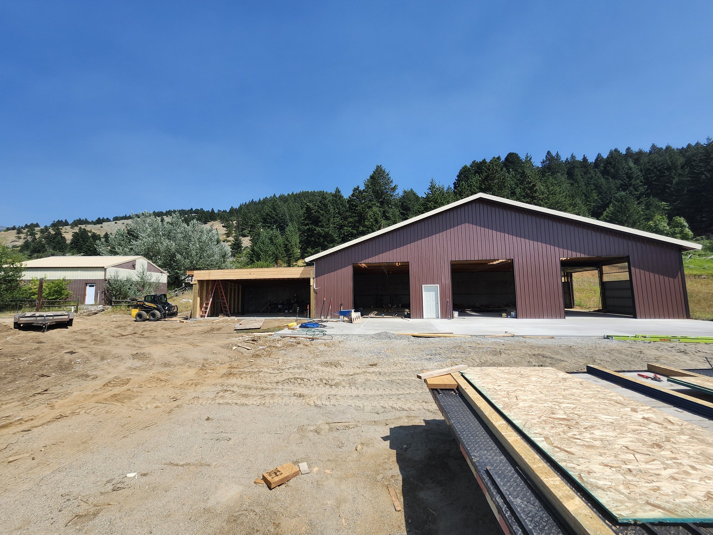 Construction site with a large brown barn-style building, dirt ground, and construction materials, with hills and trees in the background on a sunny day.