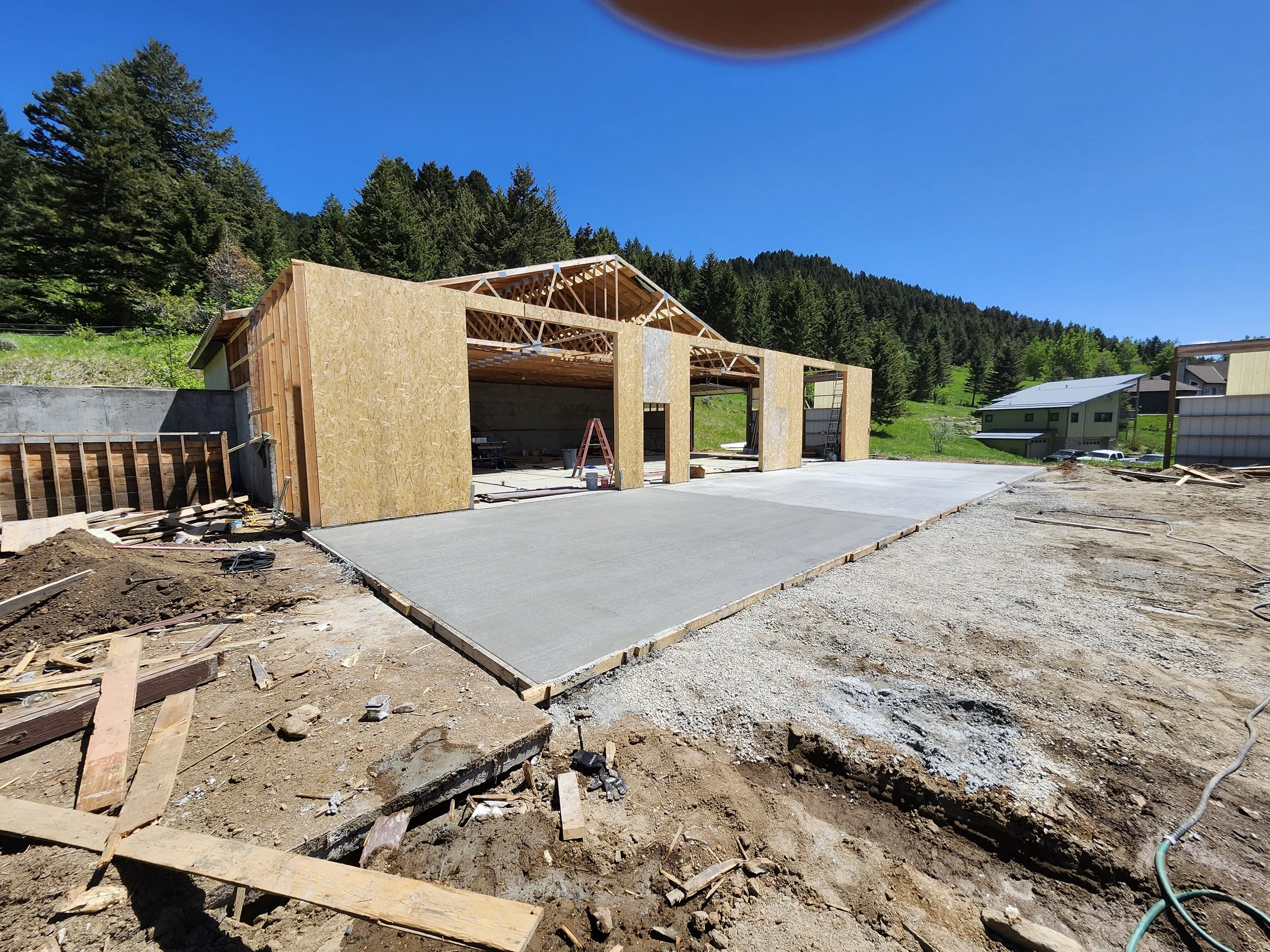 Construction site with a partially built garage or storage building, with wooden framing and concrete slab, surrounded by dirt and construction materials, amid a rural hillside with trees and neighboring houses under a clear blue sky.