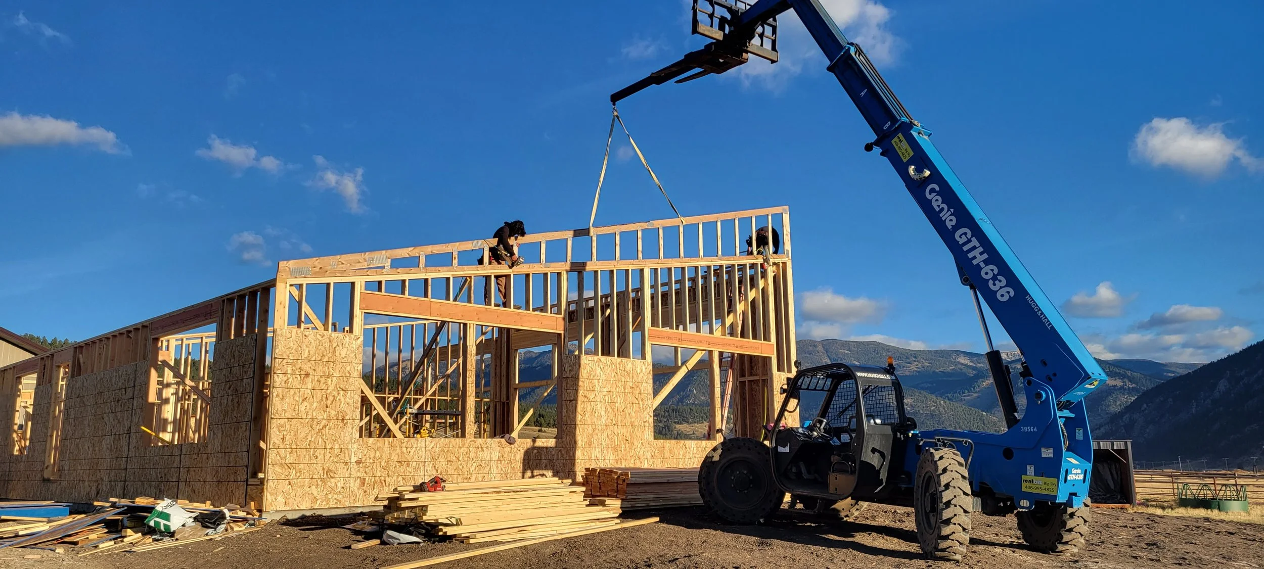 Construction workers building a wooden house frame with a crane on a construction site, mountains in the background, and a clear blue sky.