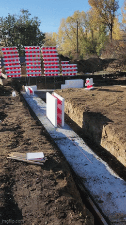 Construction site with concrete foundation outlines and red and white safety barriers, trees in the background.