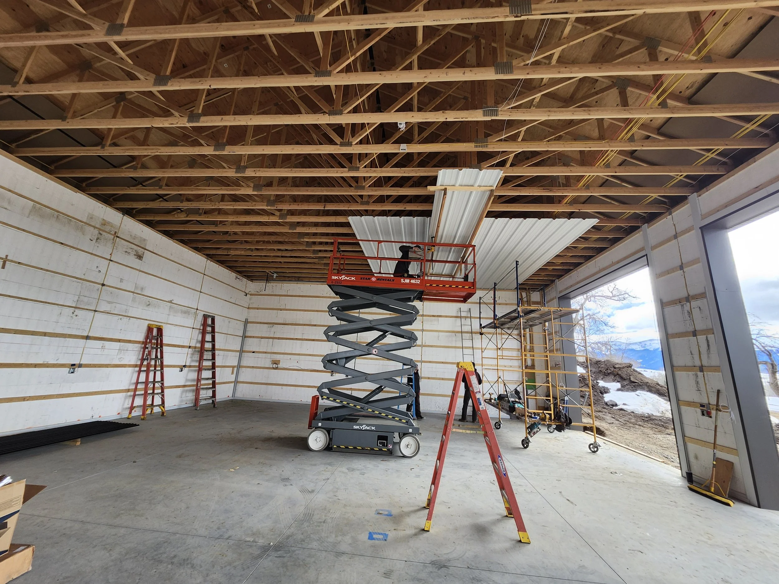 Construction workers installing ceiling panels in an unfinished building with exposed wooden roof trusses, scaffolding, a cherry picker lift, ladders, and large open windows showing a mountain landscape.