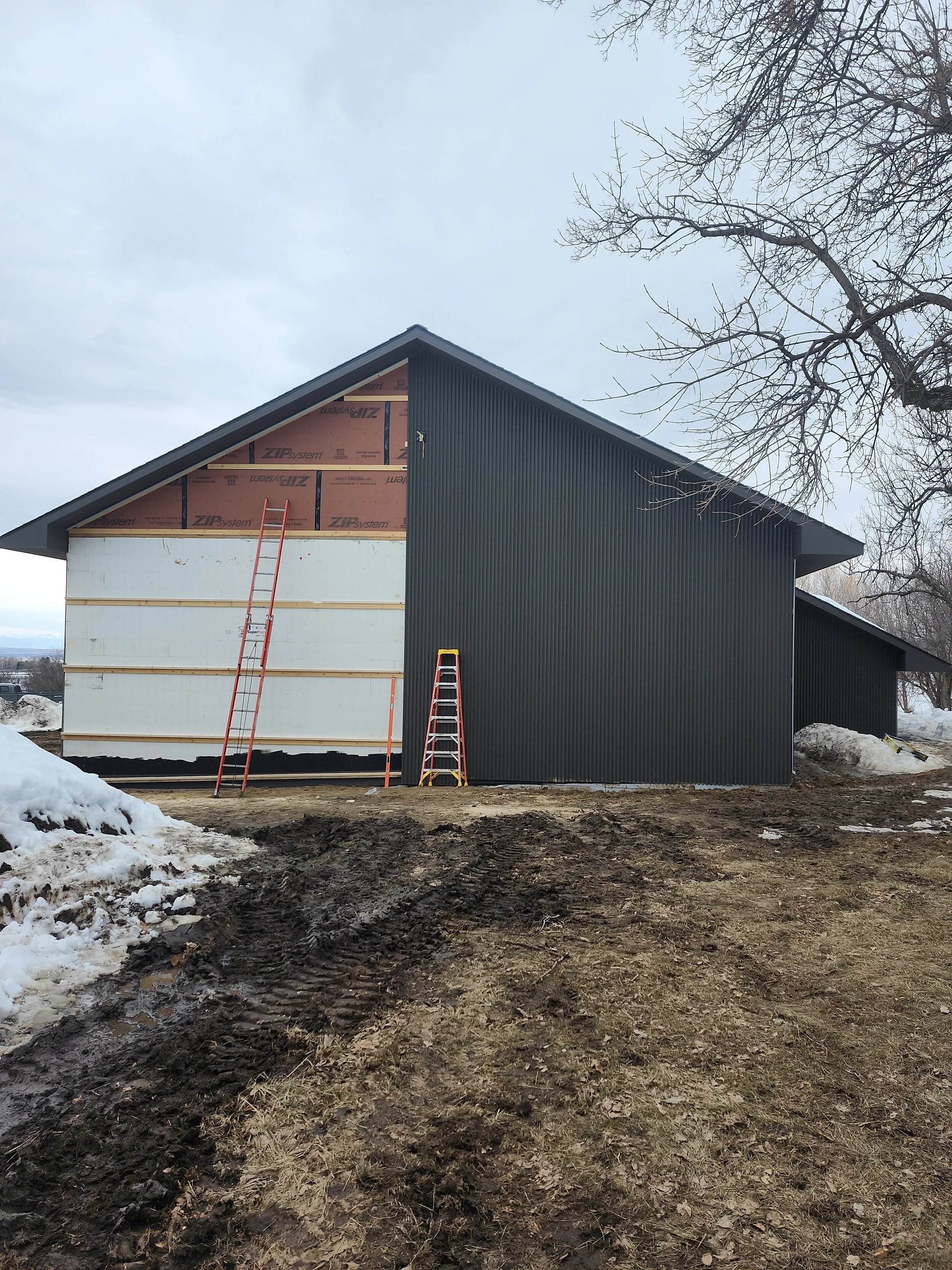 A house under construction with gray siding, a sloped roof, and snow on the ground. Two ladders lean against the house, and an unfinished section reveals insulation and construction materials.