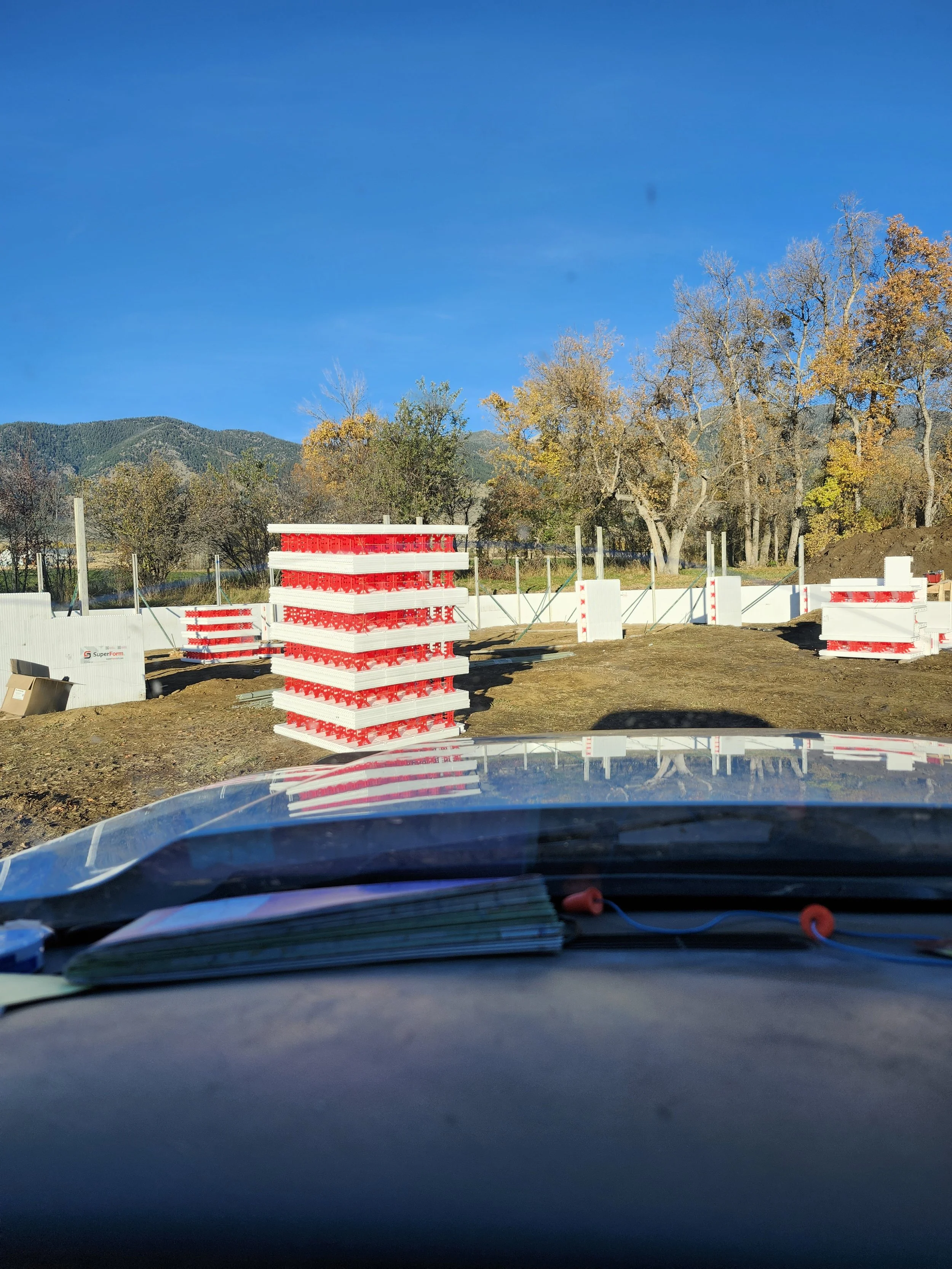 View from inside a vehicle shows a construction site with temporary white and red barriers, a tall stack of white and red construction materials, and trees with autumn foliage against a clear blue sky. Mountainous terrain is visible in the background.