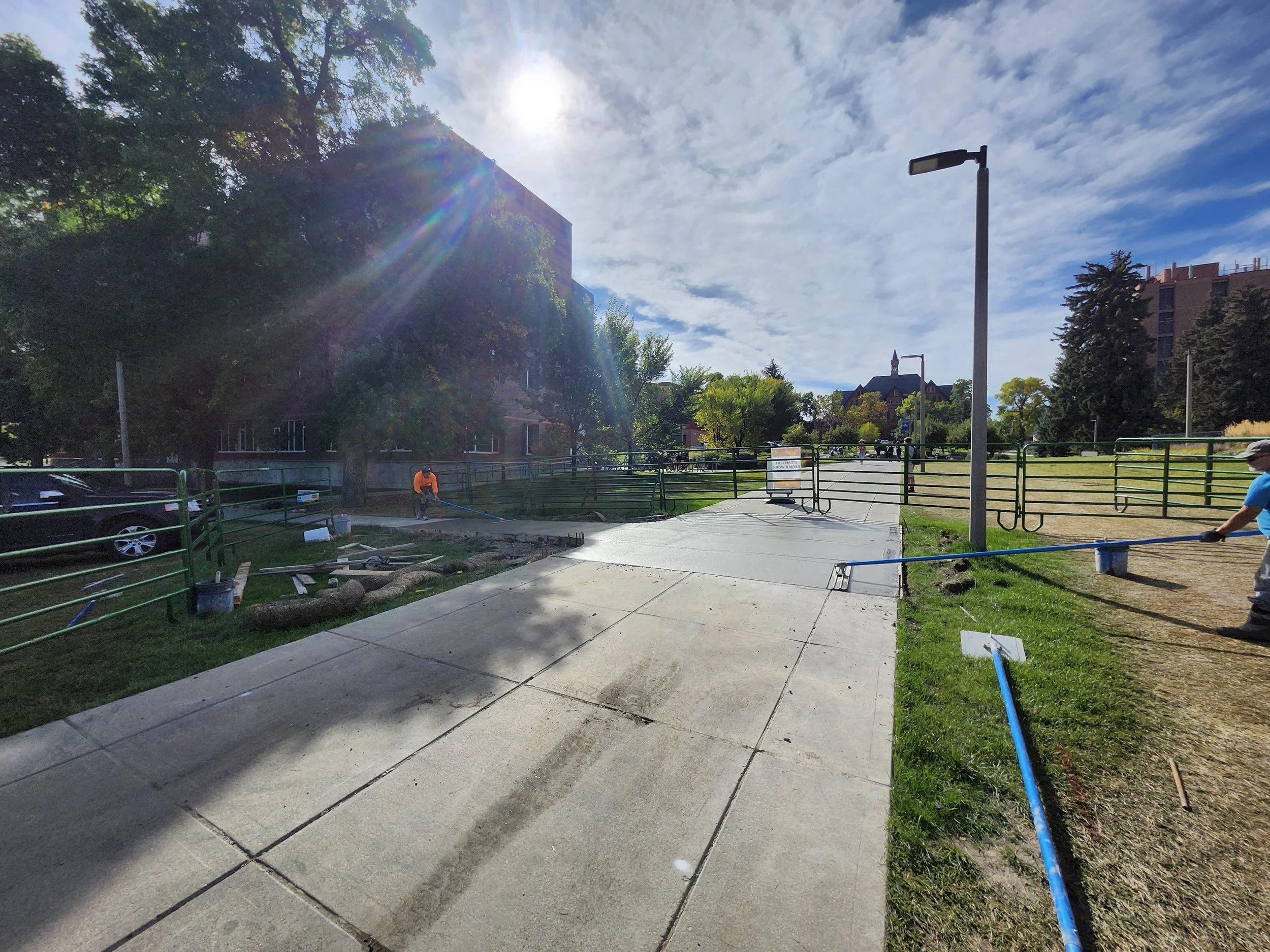Workers are repairing or resurfacing a sidewalk in a park or urban area, with some construction equipment present. The scene is sunny with a partly cloudy sky, trees, and buildings in the background.