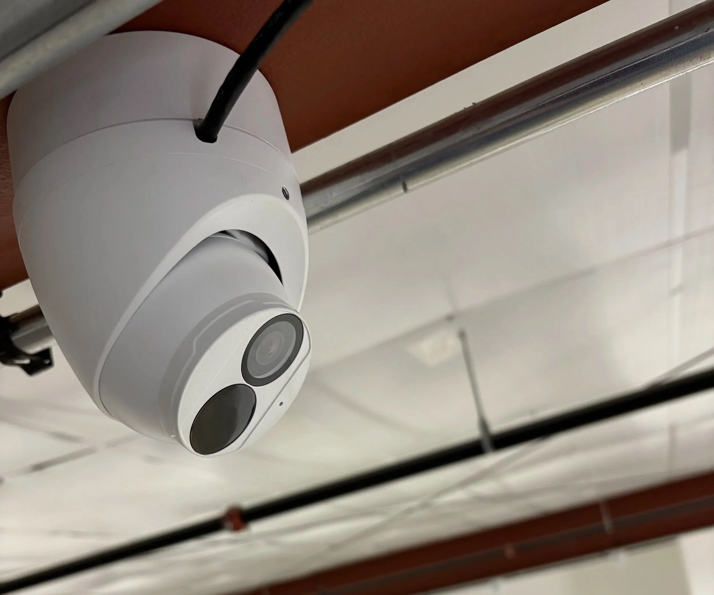 A white security camera mounted on a ceiling with pipes and ceiling tiles visible in the background.