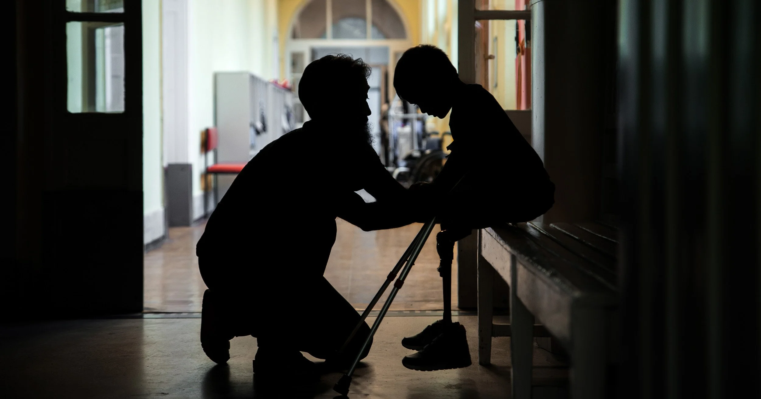 Silhouetted caregiver assisting a seated person with a walker in a hallway