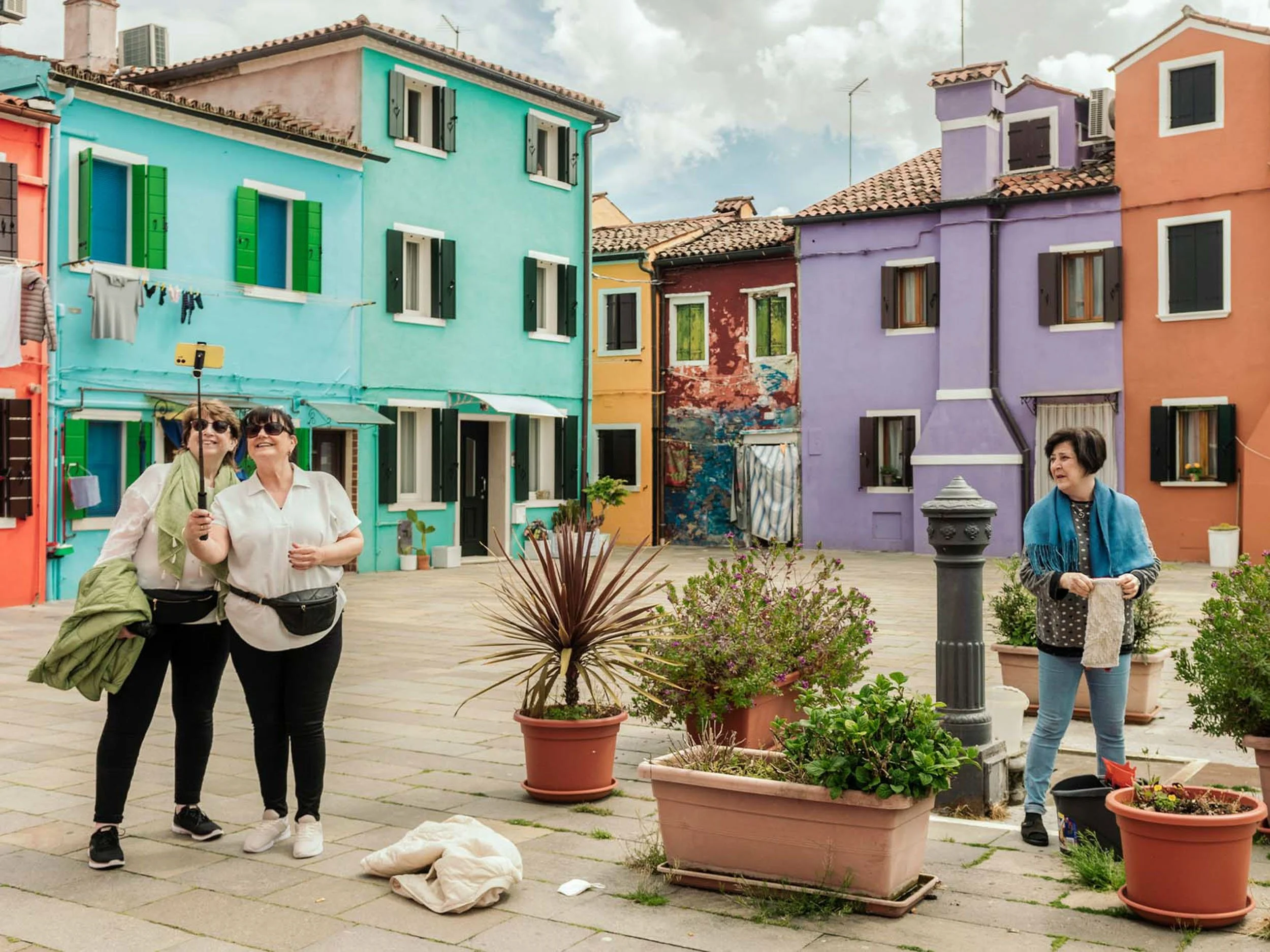 Tourists in a colorful Venetian square surrounded by brightly painted houses, potted plants, and a historic wellhead
