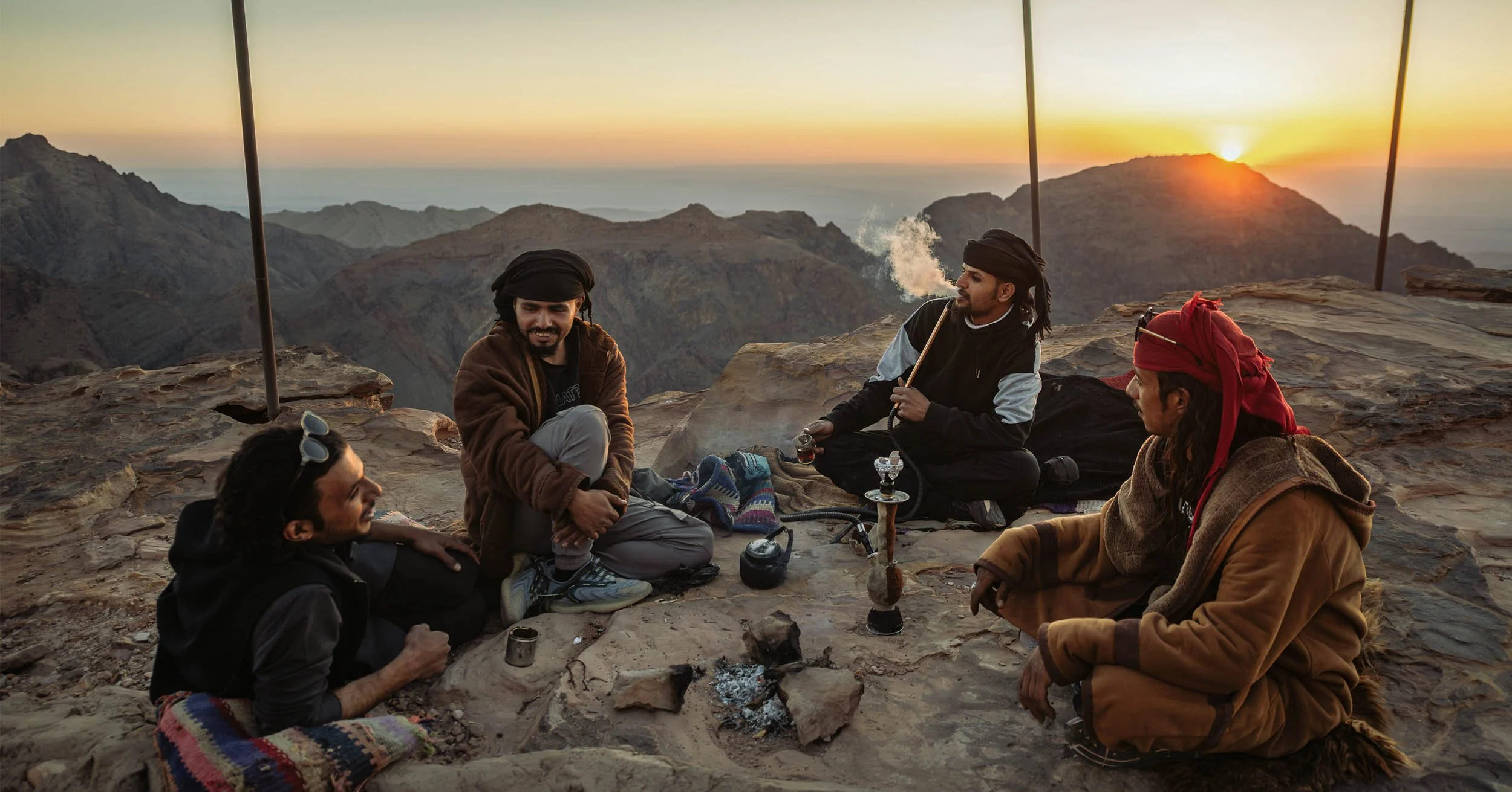 Four men in traditional clothing gather around a hookah on a rocky mountain overlook at sunset.