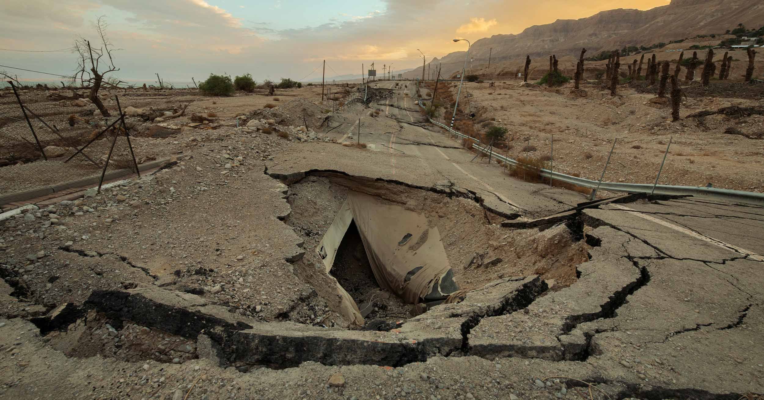 Severely cracked road with a collapsed drainage culvert in a barren desert landscape beside the Dead Sea and mountains at sunset