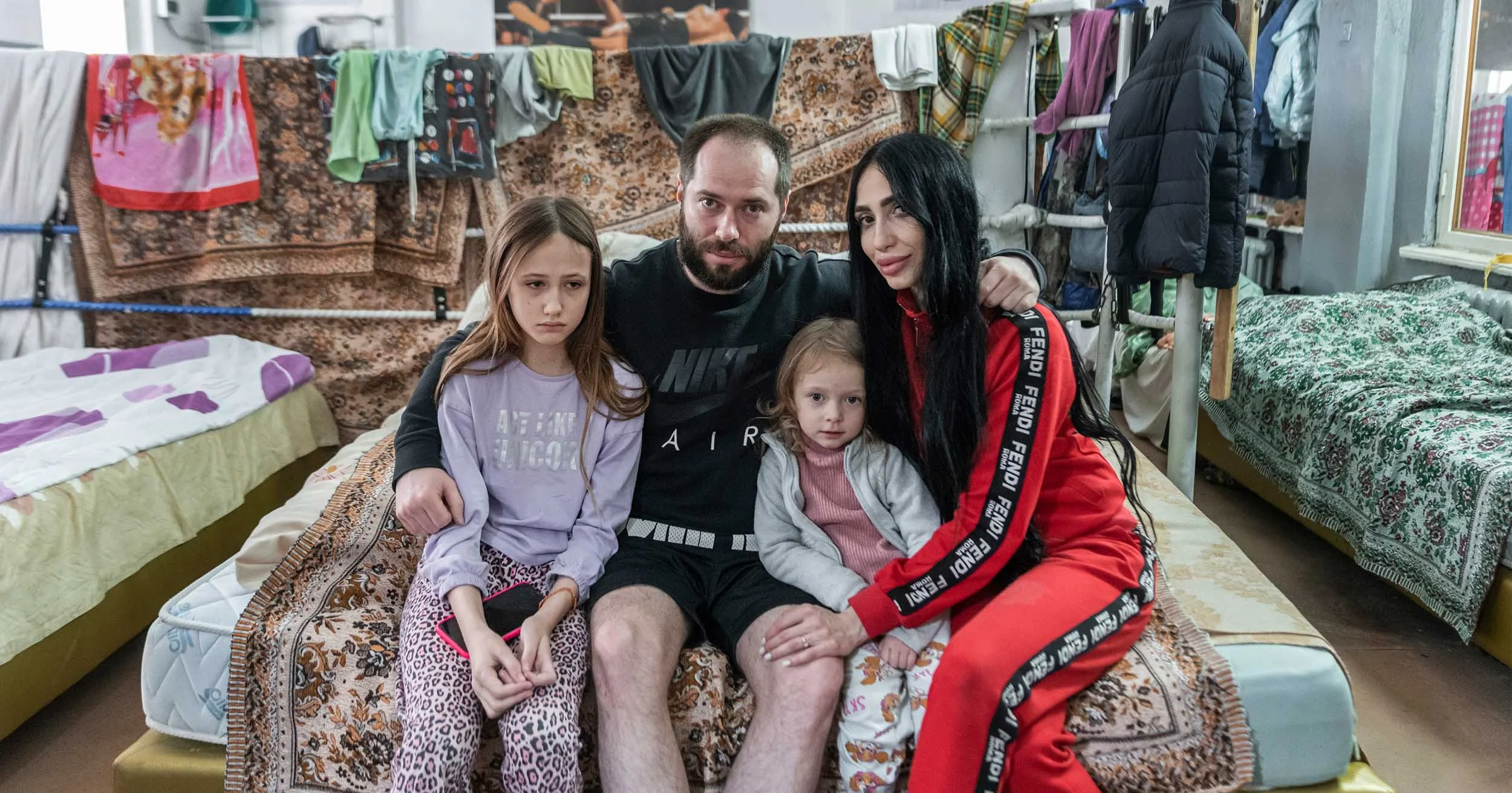 Family of four posing together on a bed in a crowded room with clothes and blankets hanging around them