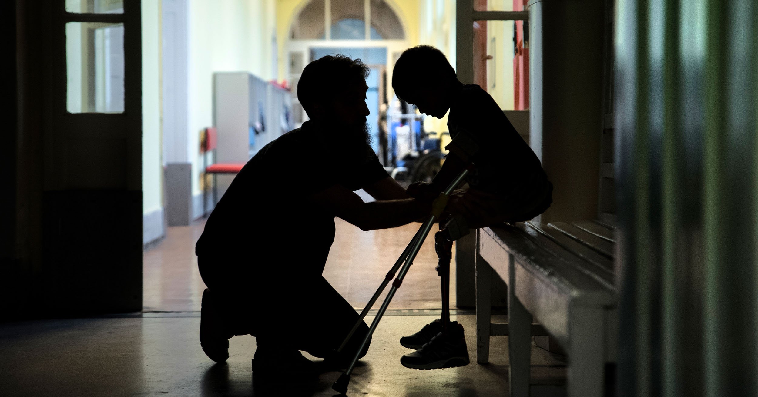 Silhouetted caregiver assisting a seated person with a walker in a hallway