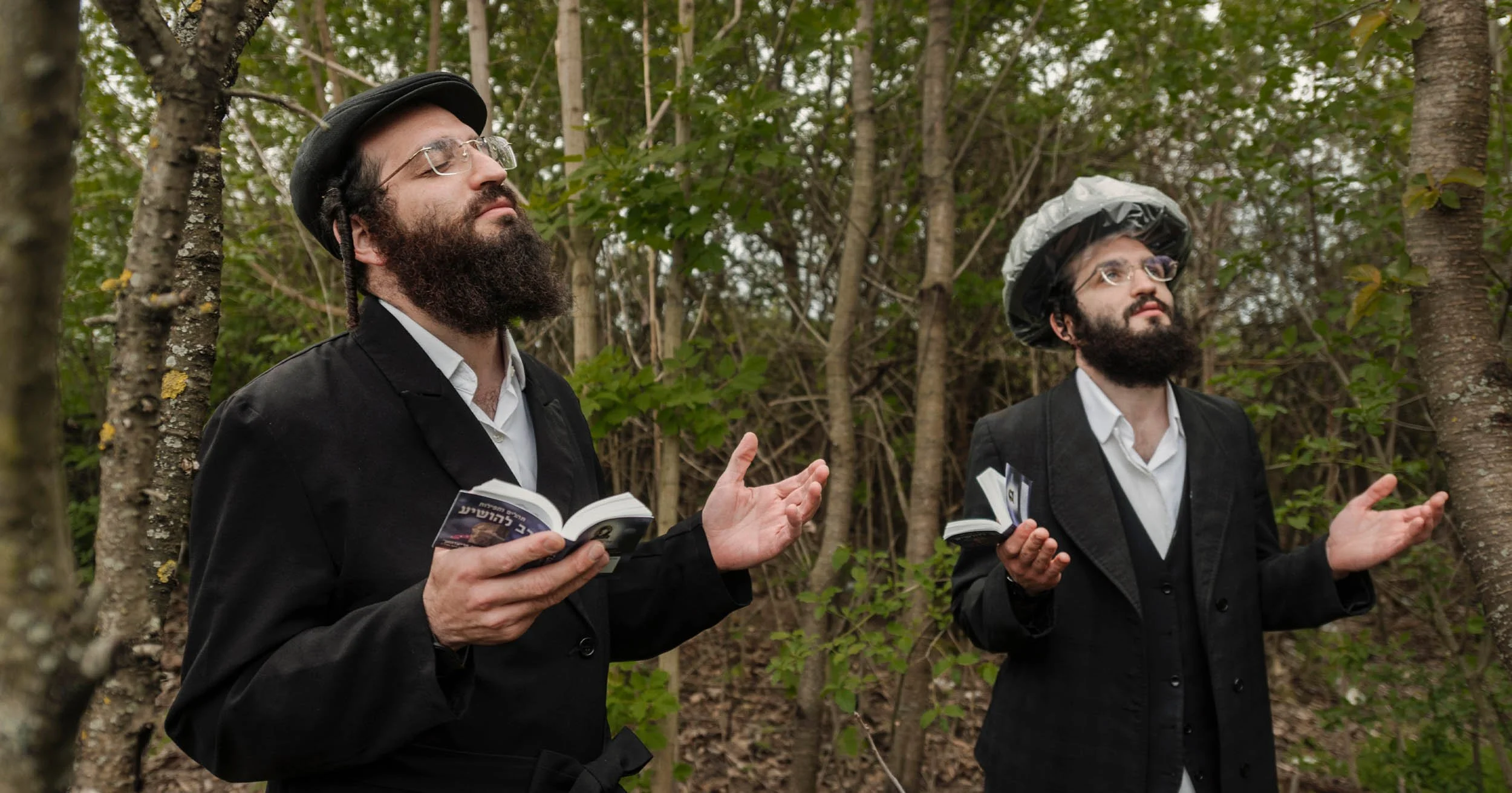 Two bearded men in black suits and hats read books and gesture in a forest.