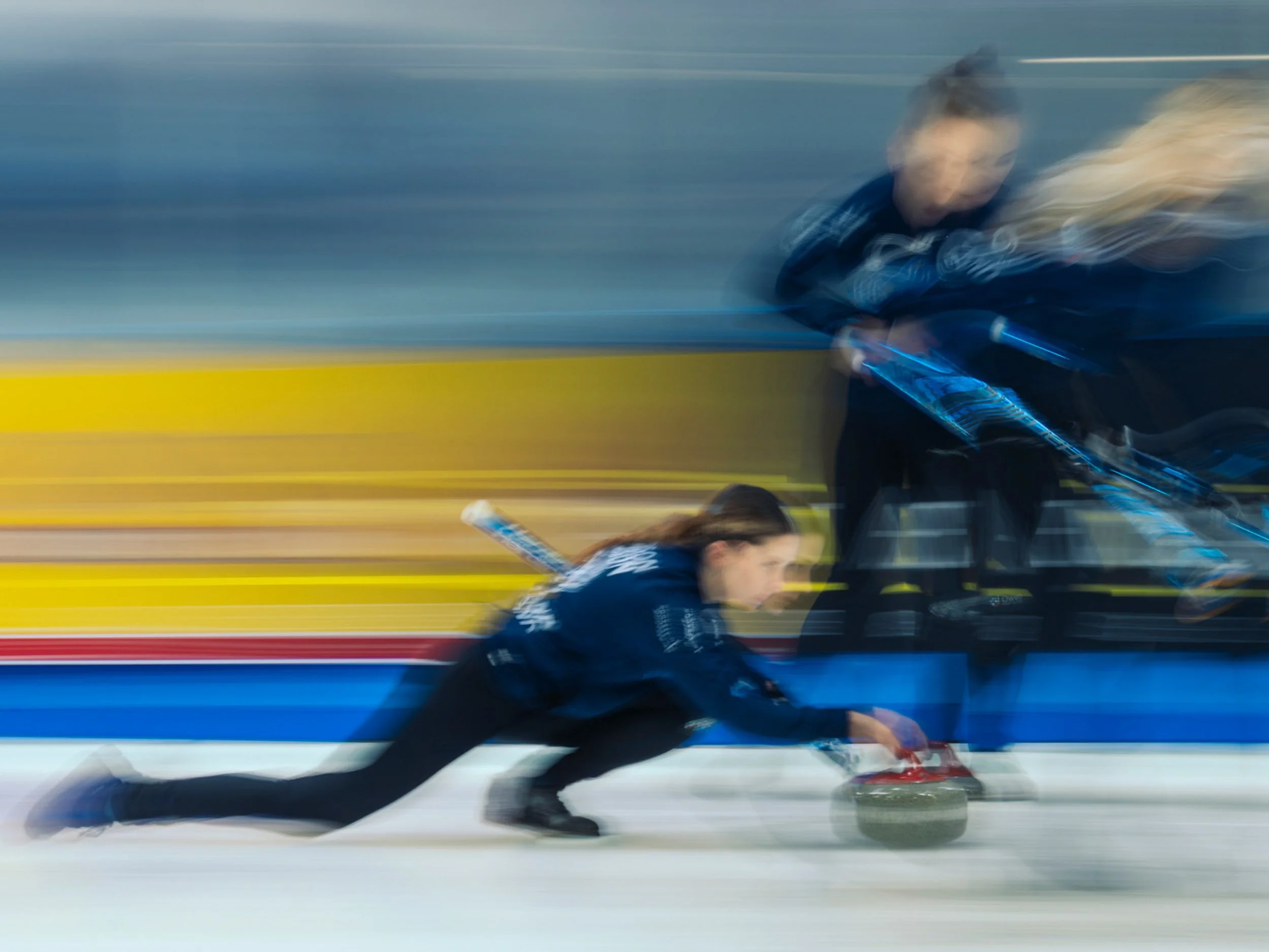 Motion-blurred female curlers sweeping ice in front of a curling stone during a match