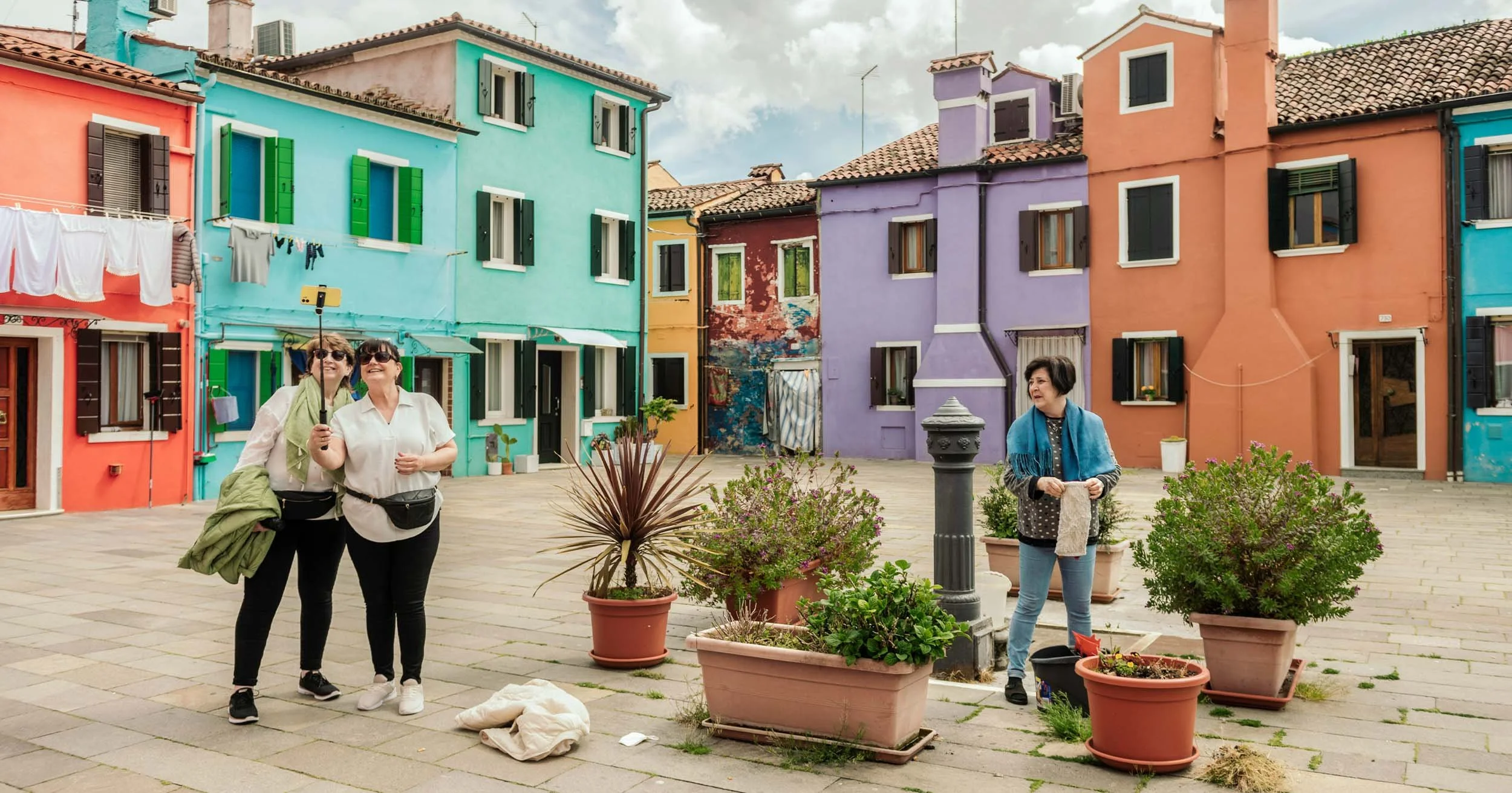 Tourists in a colorful Venetian square surrounded by brightly painted houses, potted plants, and a historic wellhead