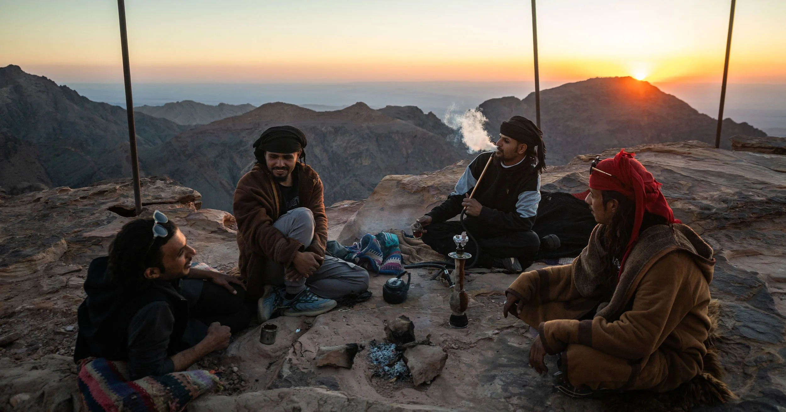 Four men in traditional clothing gather around a hookah on a rocky mountain overlook at sunset.