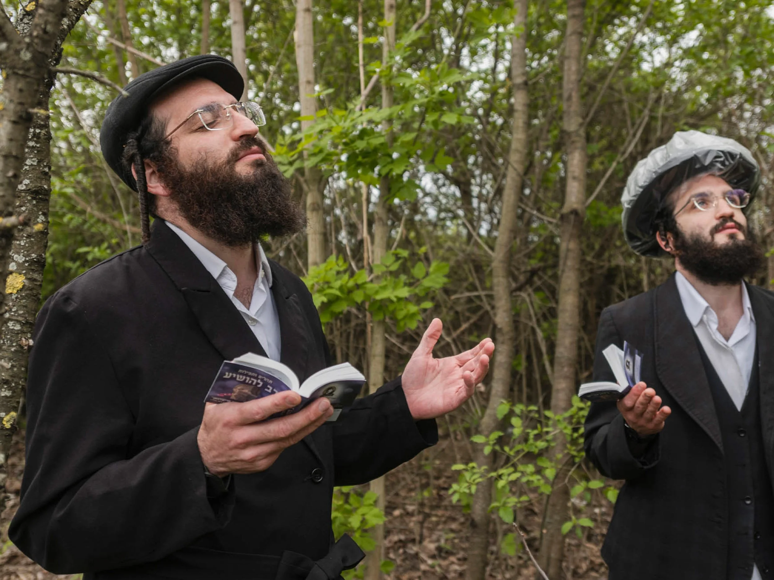 Two bearded men in black suits and hats read books and gesture in a forest.