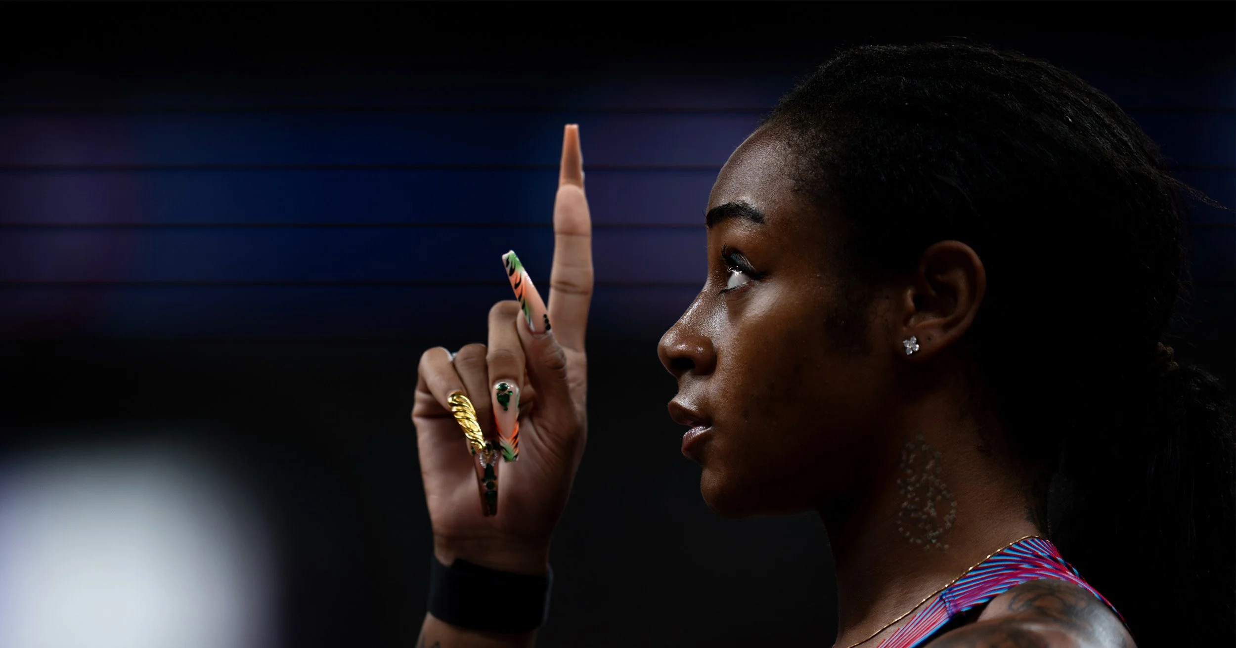 Profile portrait of a Black woman with long braided hair, colorful nails, jewelry, and raised index finger against a dark blue background