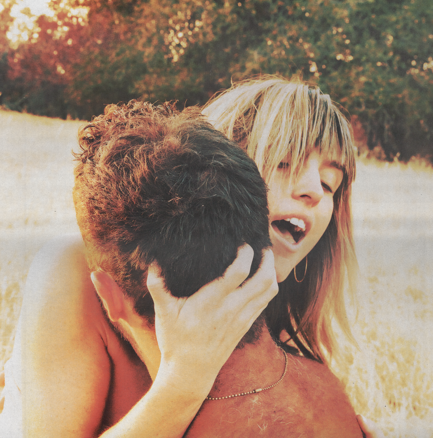 A woman with blonde hair and sunglasses holding a man with dark hair in an outdoor setting during sunset.