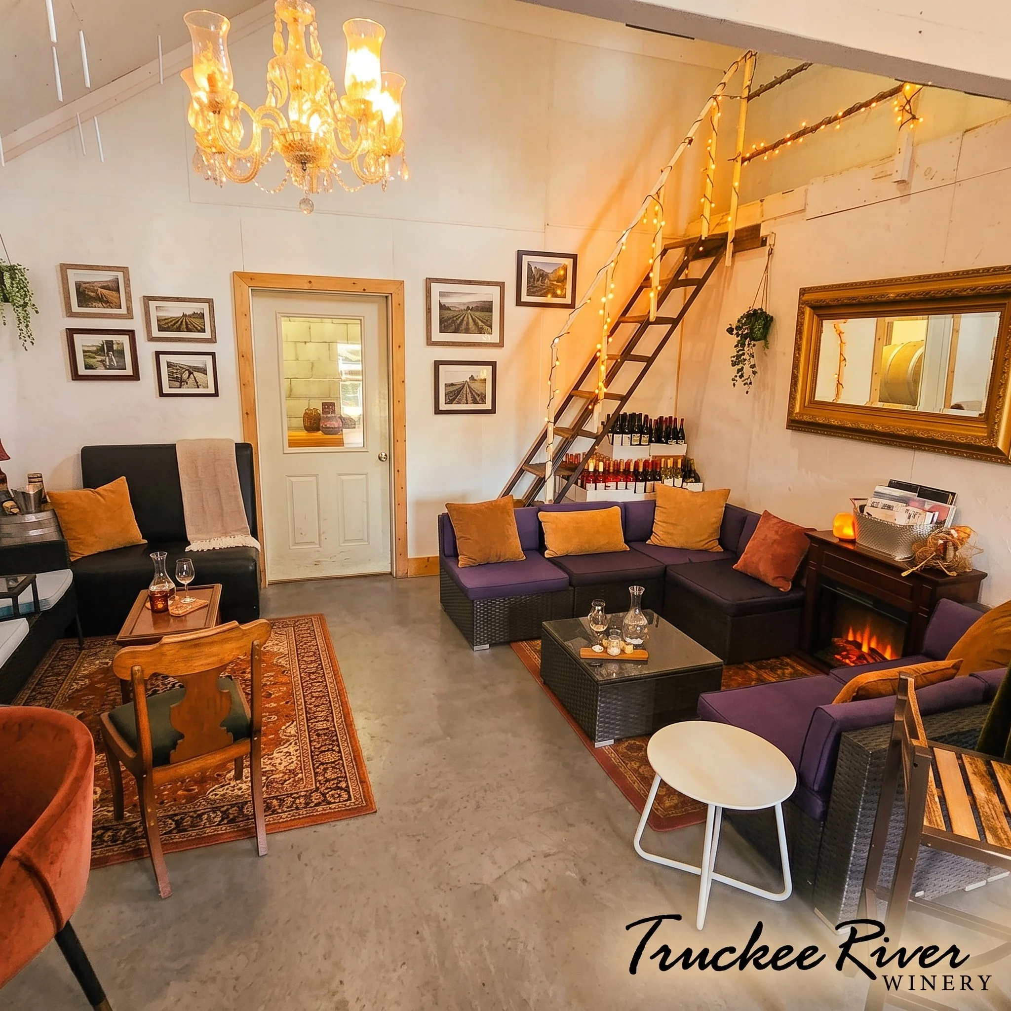 Interior of a winery with a chandelier, framed photos on the wall, a mirror, a staircase with string lights, and seating area with couches and chairs, some with pillows, and bottles of wine on a rack.