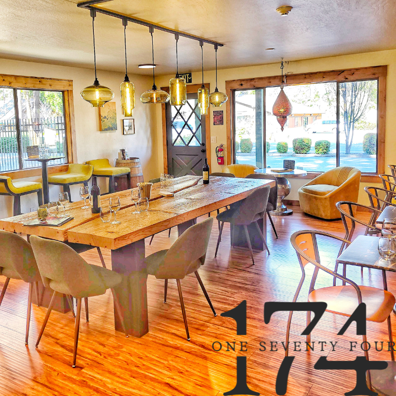 Interior of a restaurant or cafe with wooden floors, a large wooden table set with glasses and wine bottles, various seating options including chairs and armchairs, large windows letting in natural light, and decorative hanging lights.