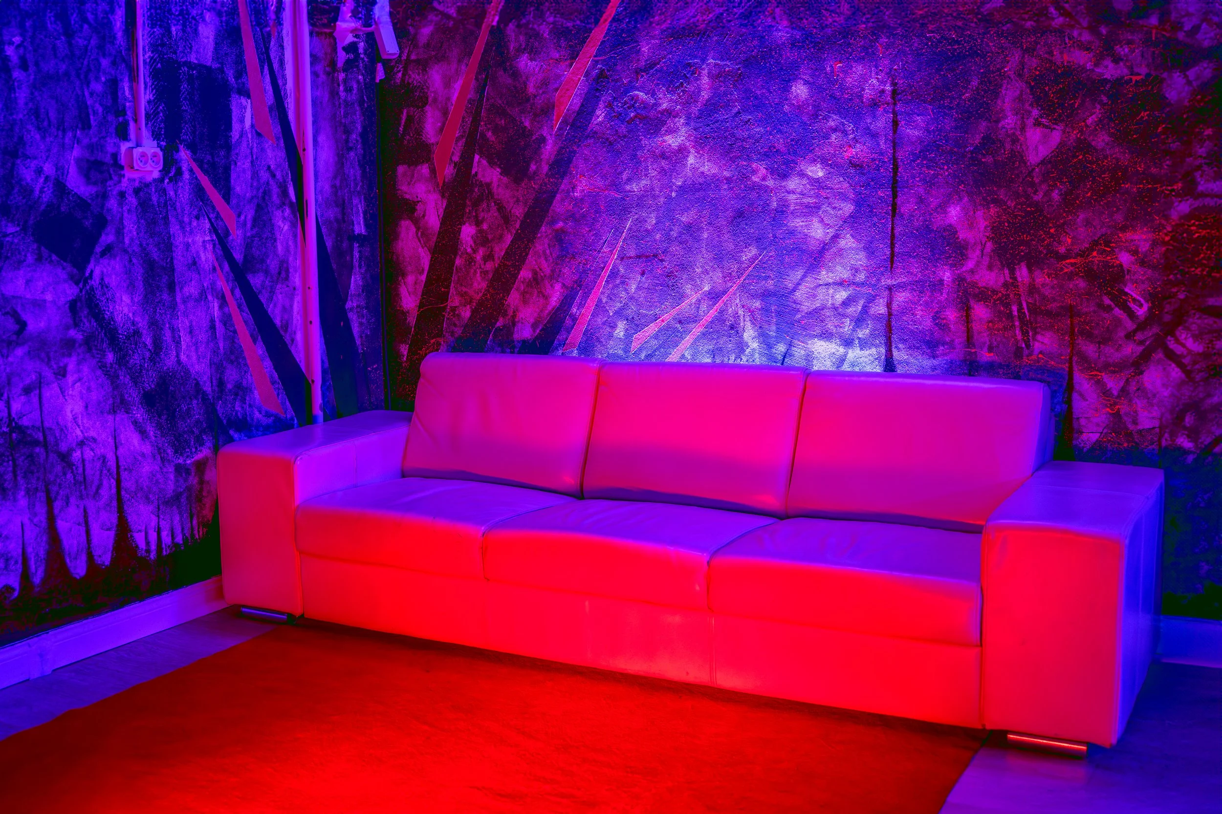 A modern pink leather sofa placed against a vibrant, multicolored, abstract wall with purple, pink, and blue hues, illuminated by colorful lighting in a room with a red carpet on the floor.