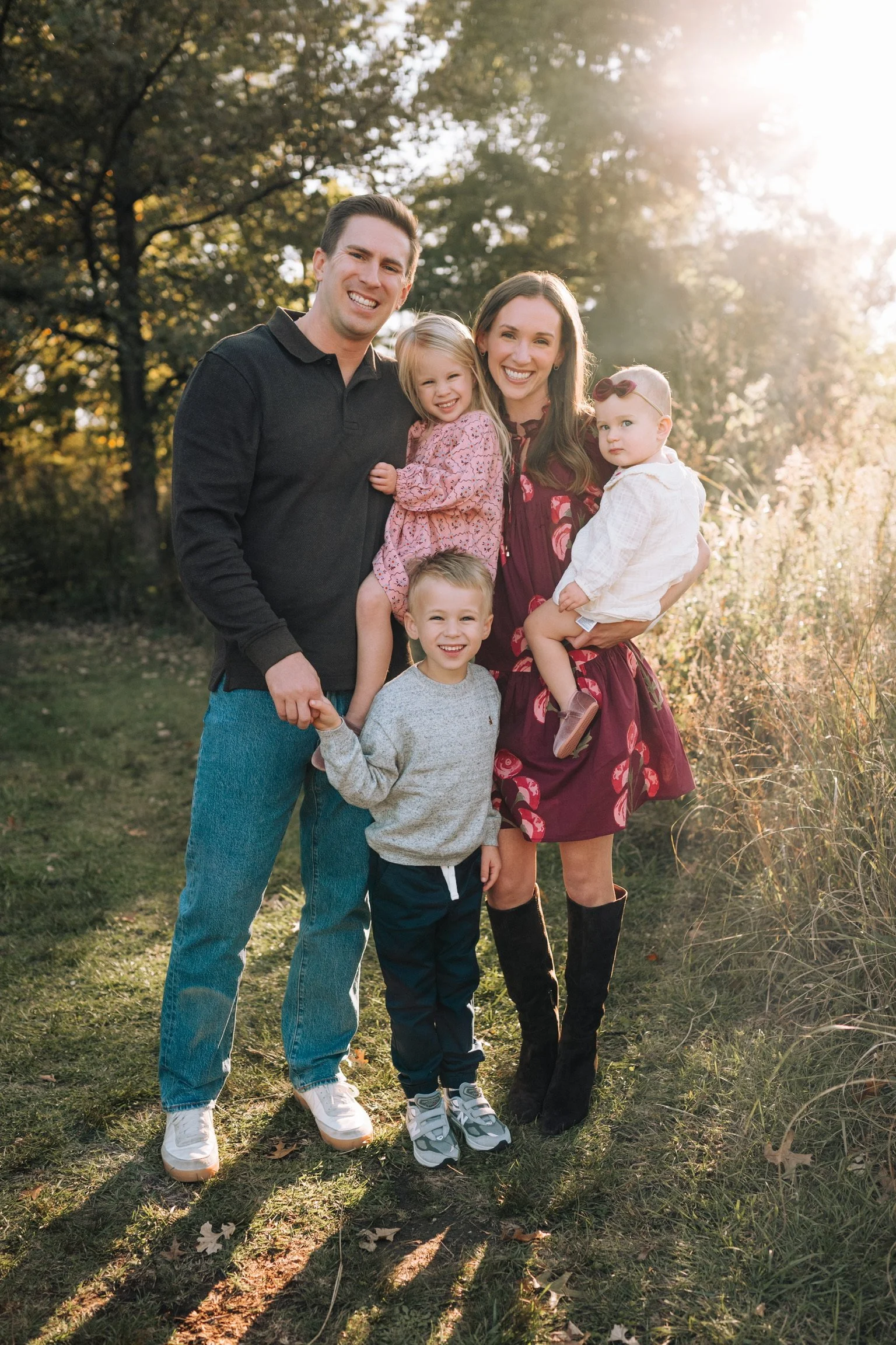 A family dressed in costumes standing on a lawn with trees and houses in the background. The woman is wearing cat ears and holding a baby, the man is dressed as an elderly person, and a young boy is dressed as a firefighter.
