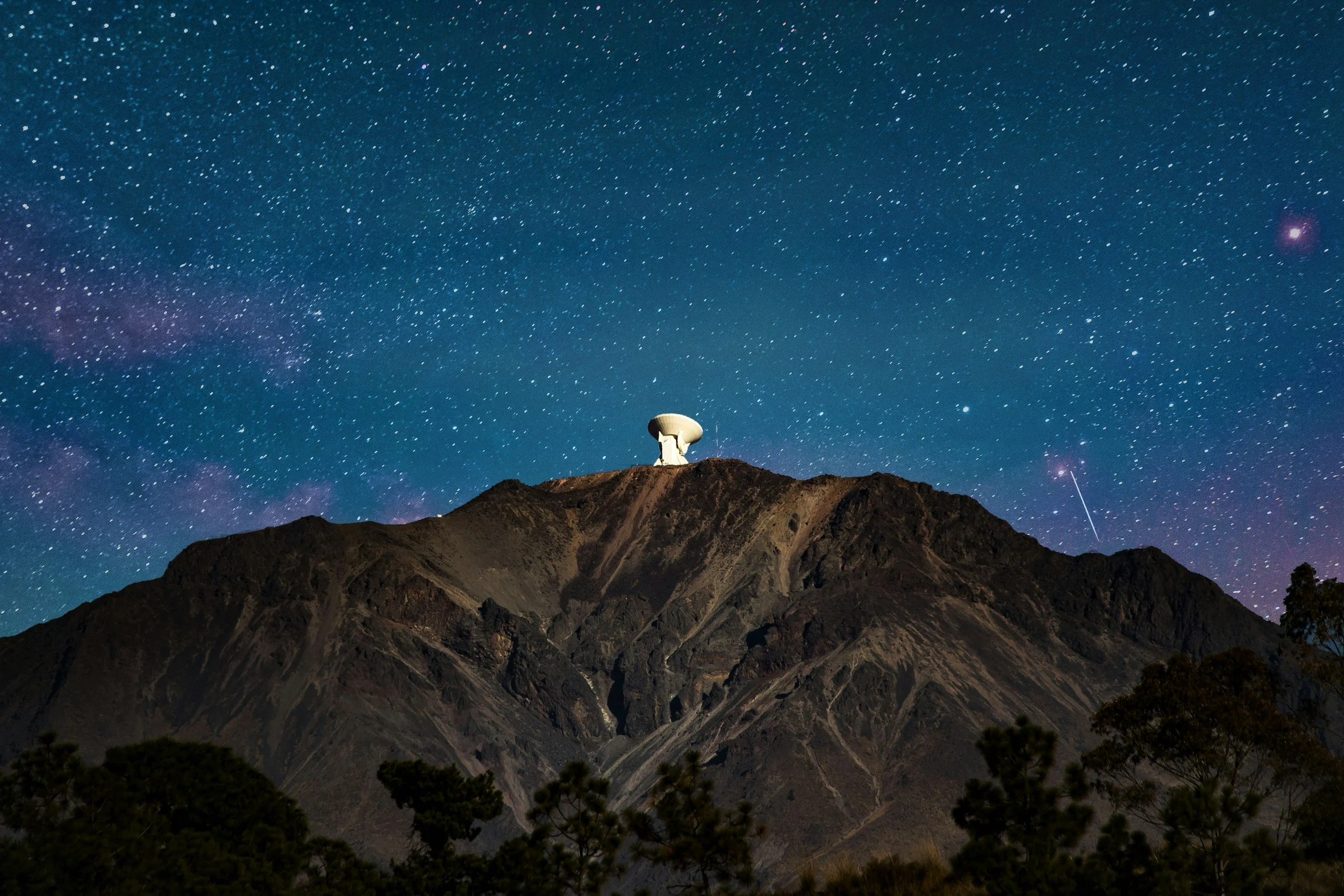 Night sky filled with numerous stars above a mountain with a satellite dish on top. Forest visible at the bottom.