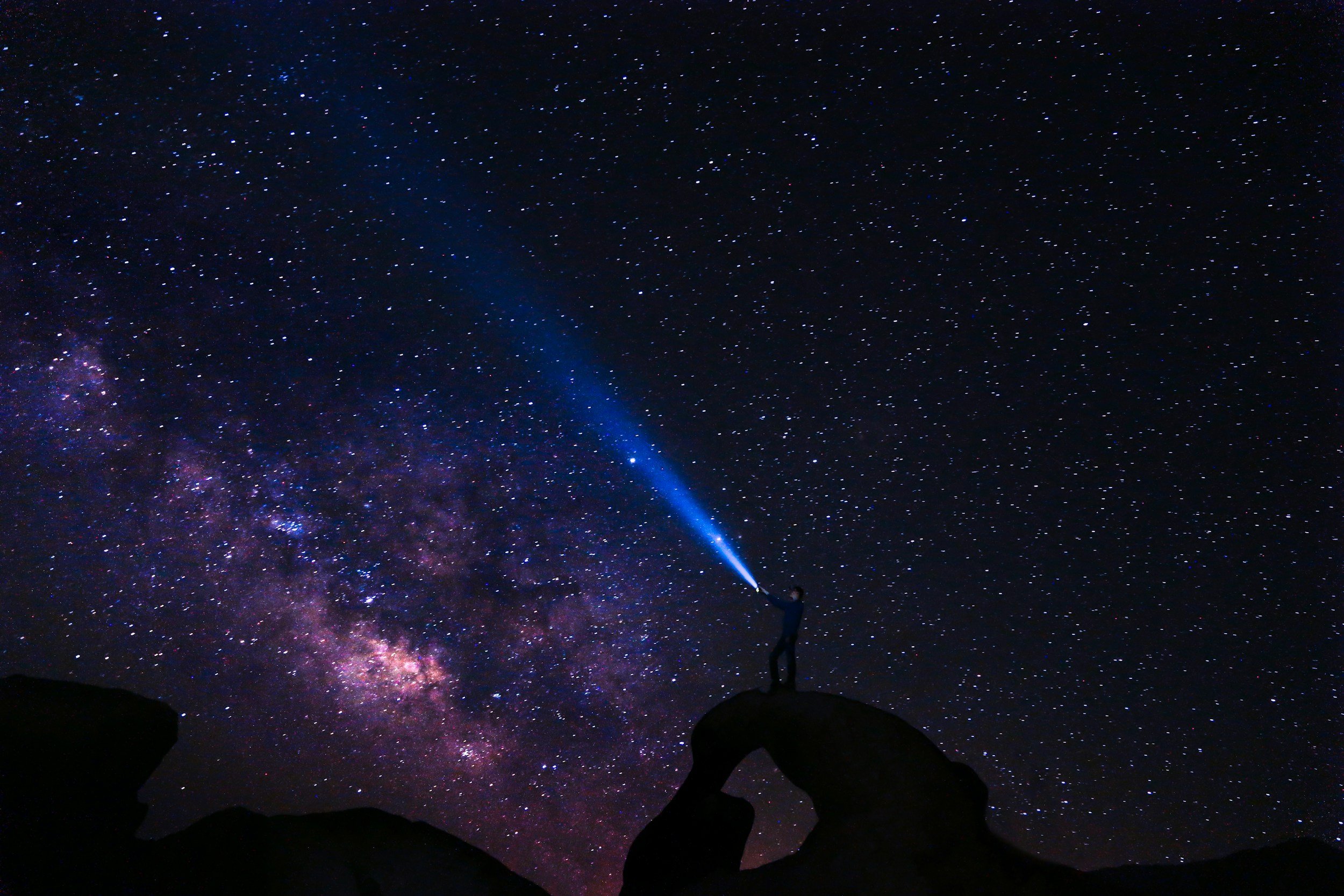 Person in silhouette standing on rocks at night, pointing a flashlight towards the starry sky and the Milky Way galaxy.